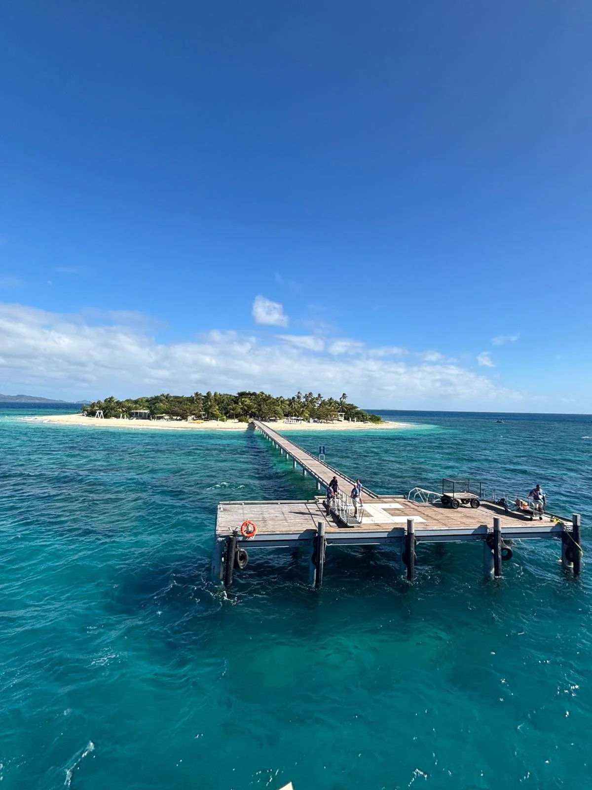 Wooden pier stretching into turquoise water toward a small tropical island with palm trees.
