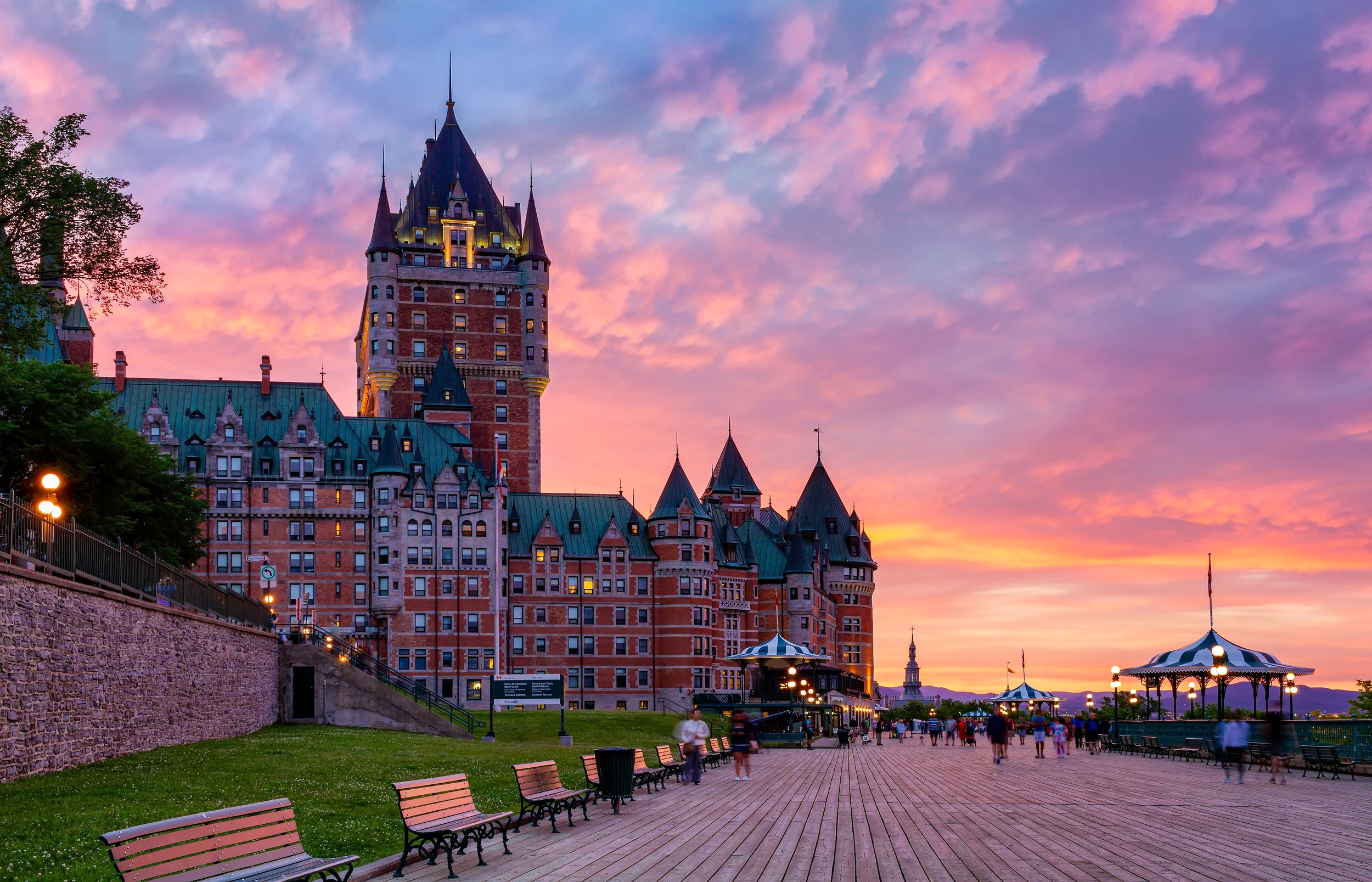 The image shows a sunset over a castle-like hotel with pointed towers and a green rooftop. In the foreground, there is a boardwalk with benches, some people walking, and a carousel with lights. The sky is filled with pink and purple clouds.