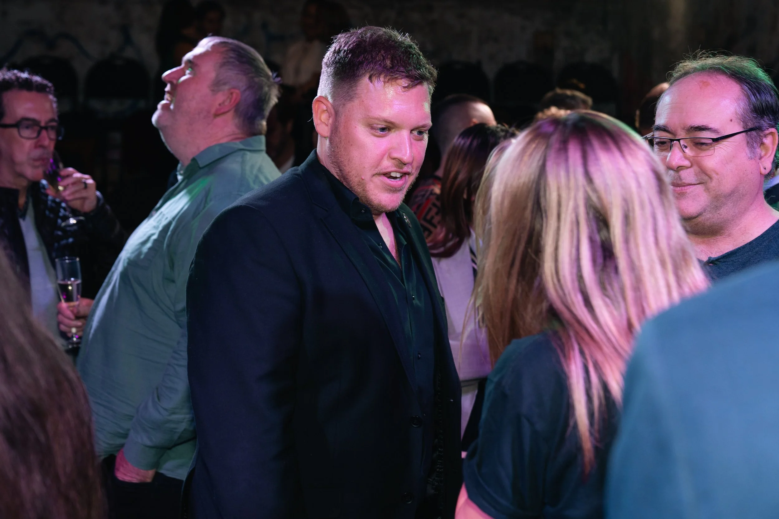 People socializing at a party, with a man in a dark shirt in the center and others around him engaged in conversations.