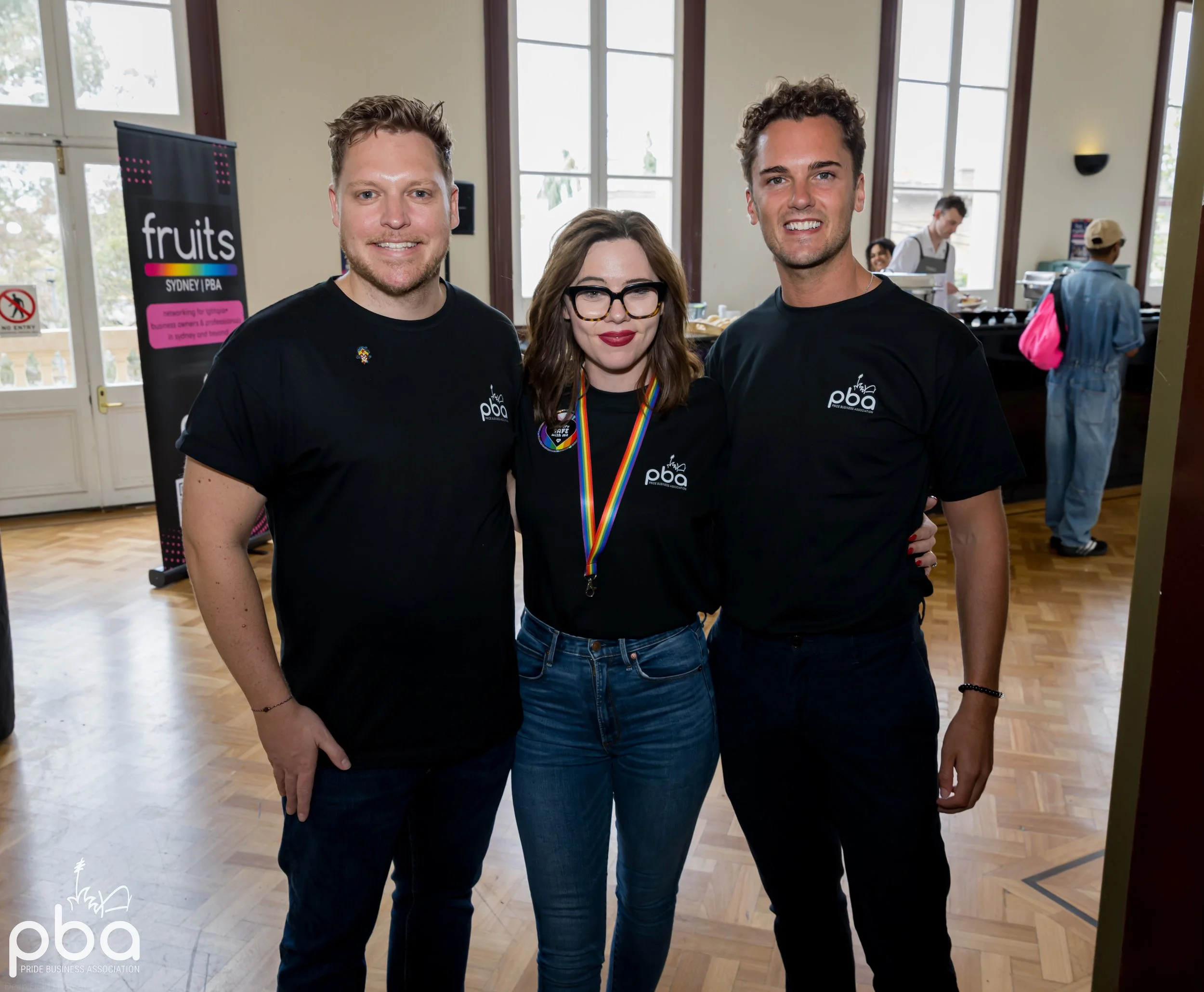 Three smiling individuals standing together indoors, with windows and other people in the background. Two men and one woman, wearing black shirts with a logo, and the woman has a rainbow-colored pride necklace.