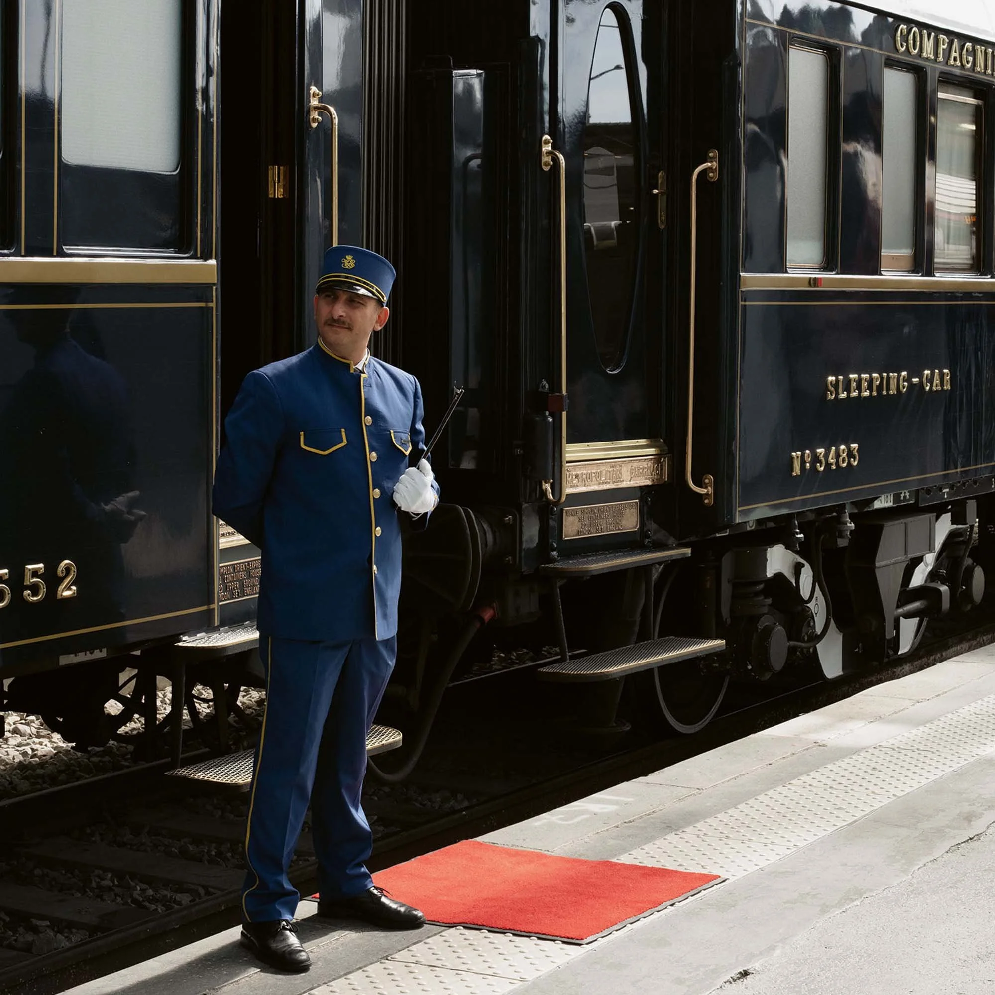 A train conductor in a blue uniform and hat standing on a platform with a red carpet, near a black historical train car.