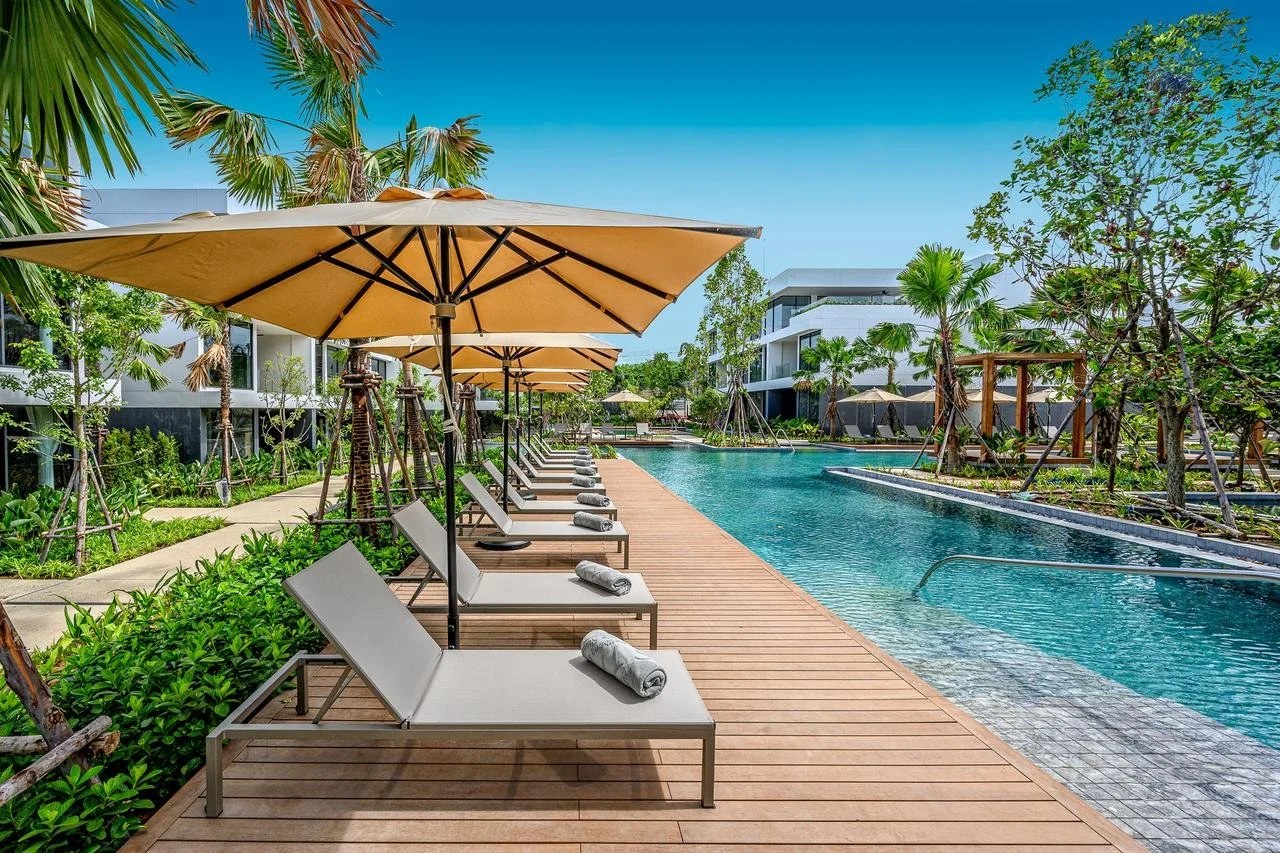 Poolside view with chairs, umbrellas, and a clear blue pool surrounded by green trees and modern white buildings under a bright blue sky.
