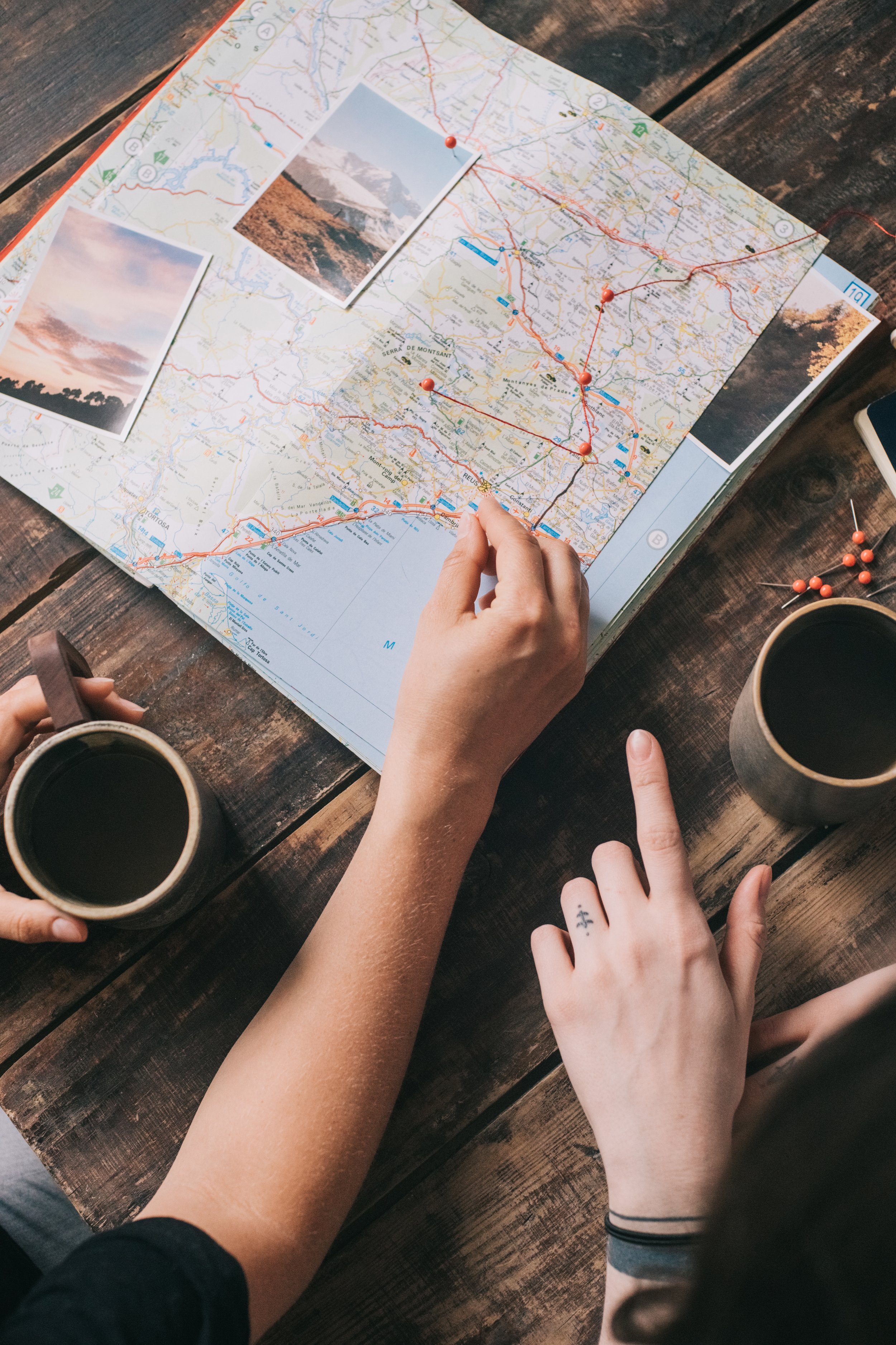 Top-down view of a map with red pins, photographs of landscapes, and hands pointing at different locations on a wooden table, with coffee mugs nearby.