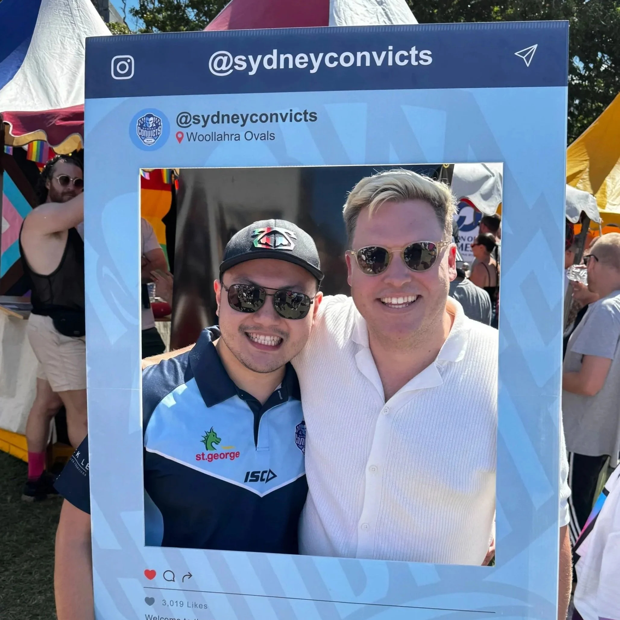 Group of people at an outdoor event taking a photo with a cardboard prop designed to look like a social media post frame, with text '@sydneyconvicts' and location 'Woollahra Ovals.' People are smiling and wearing sunglasses, with tents and trees visible in the background.