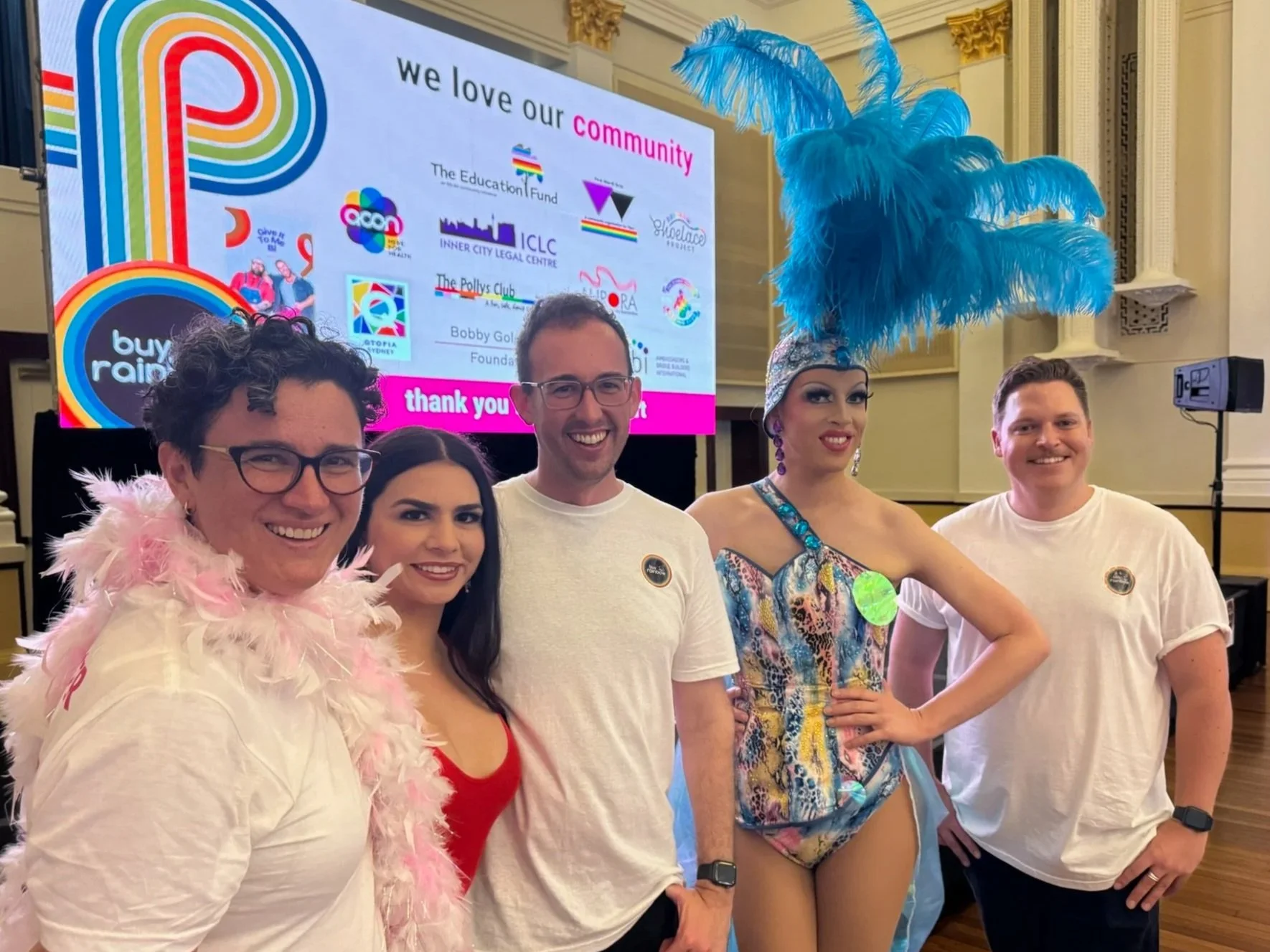 Group of five diverse individuals standing together, two women in colorful costumes and feathered headpieces, three people in casual white shirts, indoors with a large screen displaying logos and a message about supporting the community.