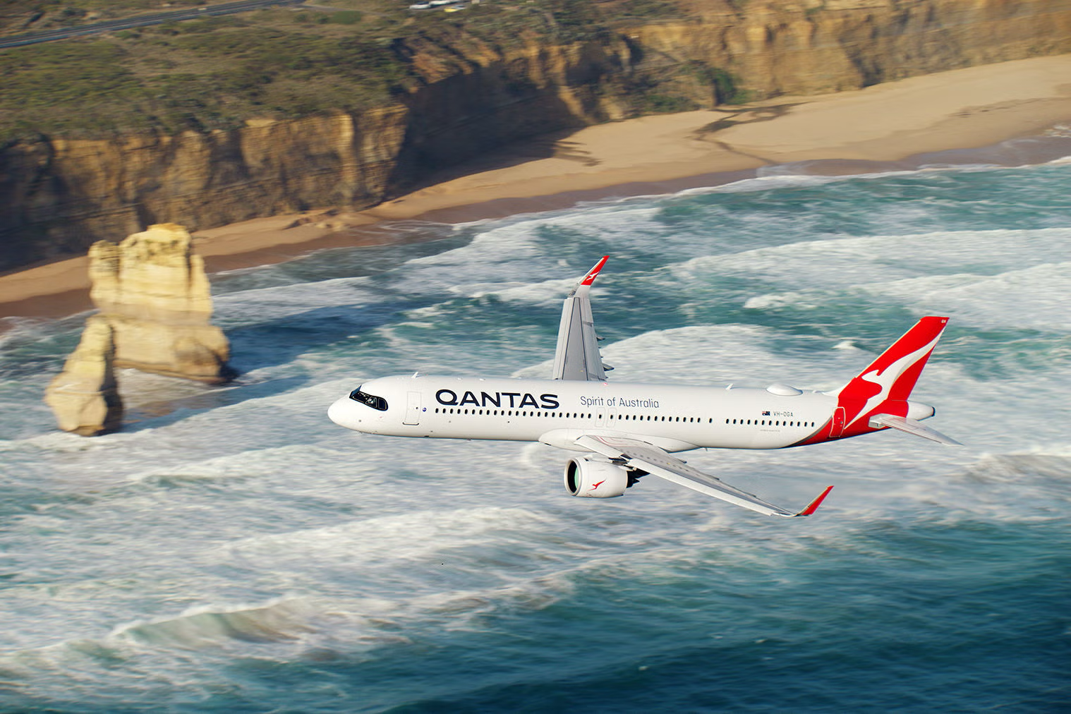 A Qantas airplane flying over the ocean near a rocky shoreline with waves crashing below.