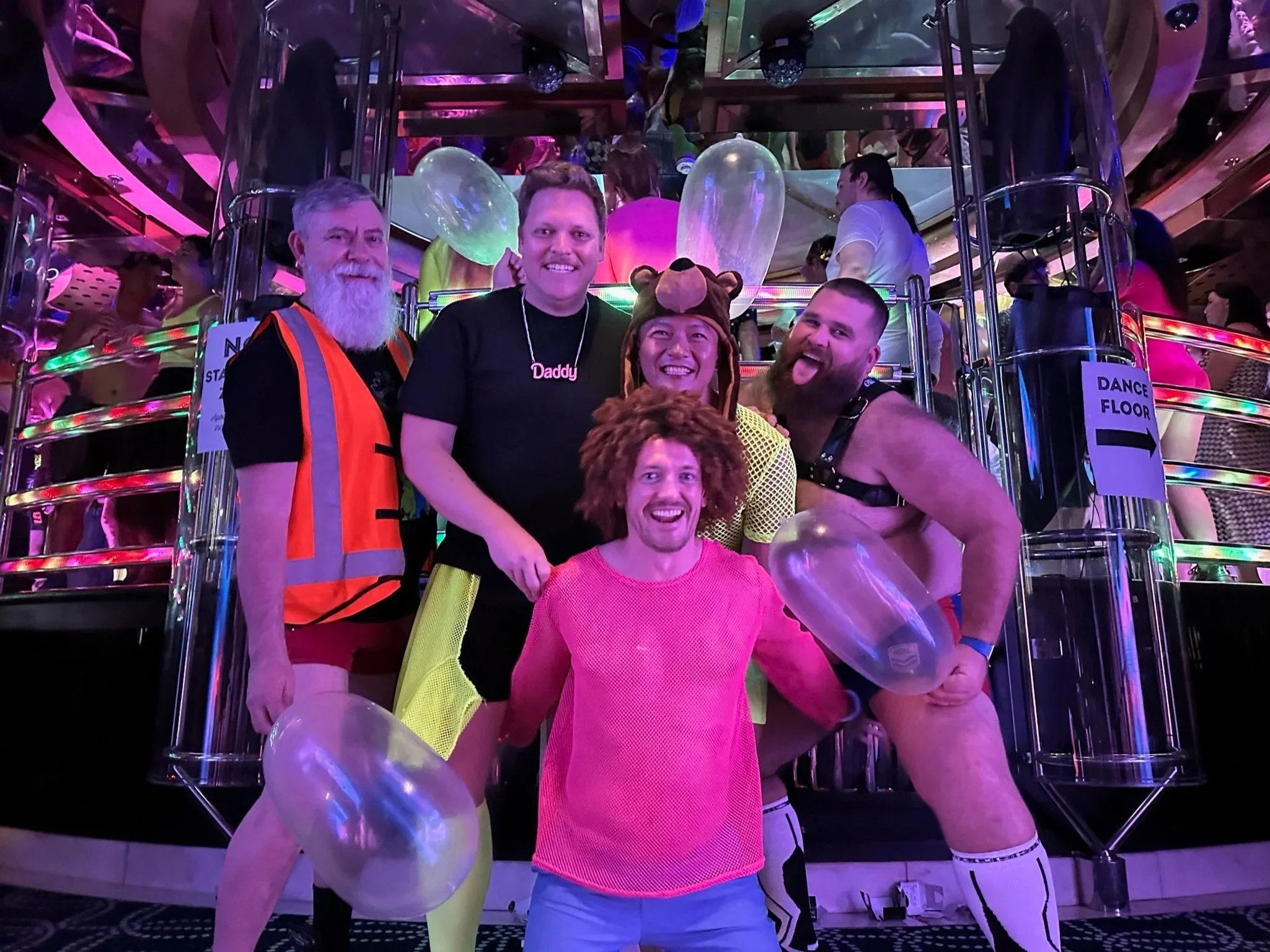 A group of five people smiling and posing for a photo at a party or event, with a colorful background including balloons, balloons, and vibrant lights. They are standing close together, with some wearing casual outfits and one person wearing a bear hat. There are other party-goers and a mirrored ceiling in the background.