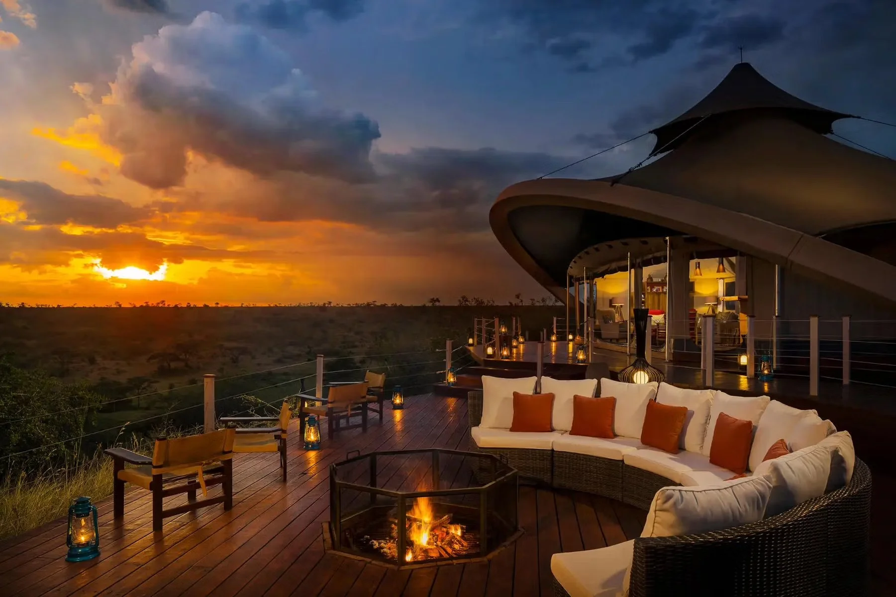 Sunset over a vast open landscape viewed from an outdoor lounge area with a modern, curved roof structure. The lounge features a sofa with orange pillows, chairs, wooden decking, and lanterns, with a small fire pit in the center casting a warm glow.