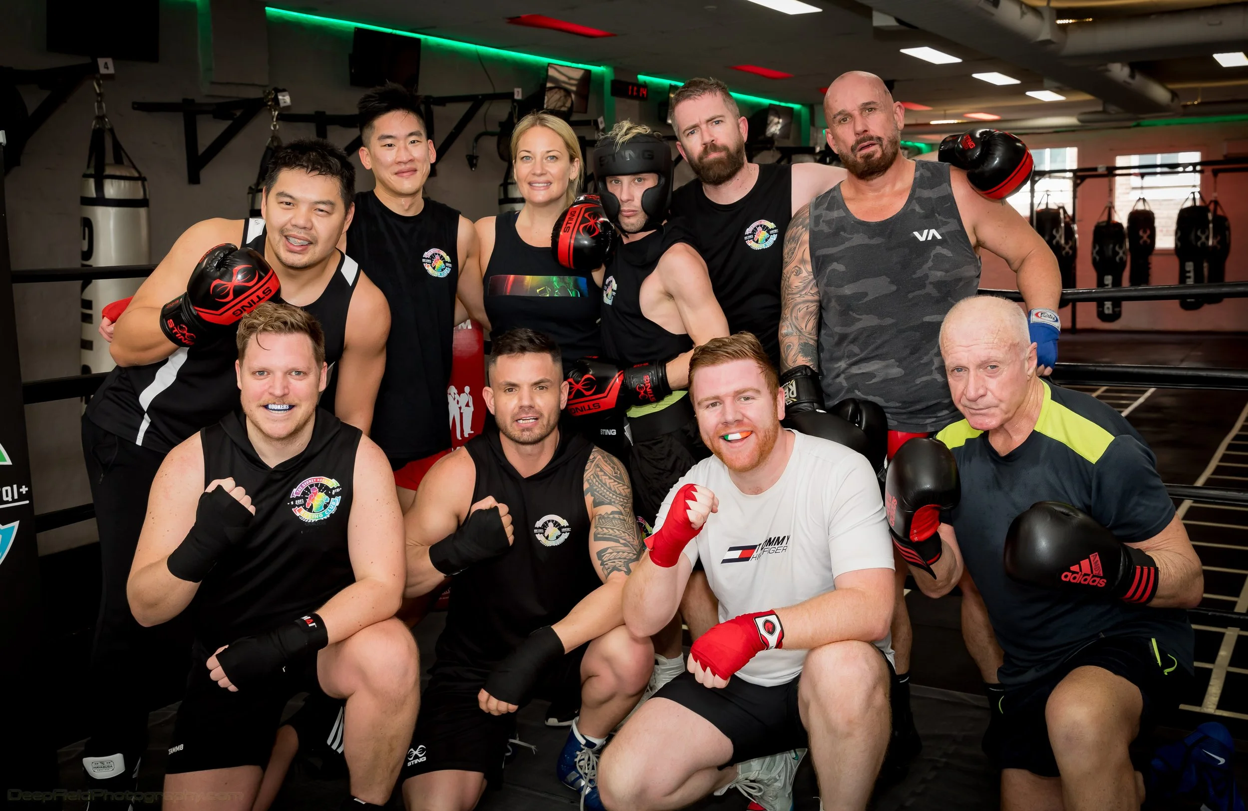 A group of ten people in a boxing gym, wearing athletic clothing and boxing gloves, posing together for a photo after a workout.