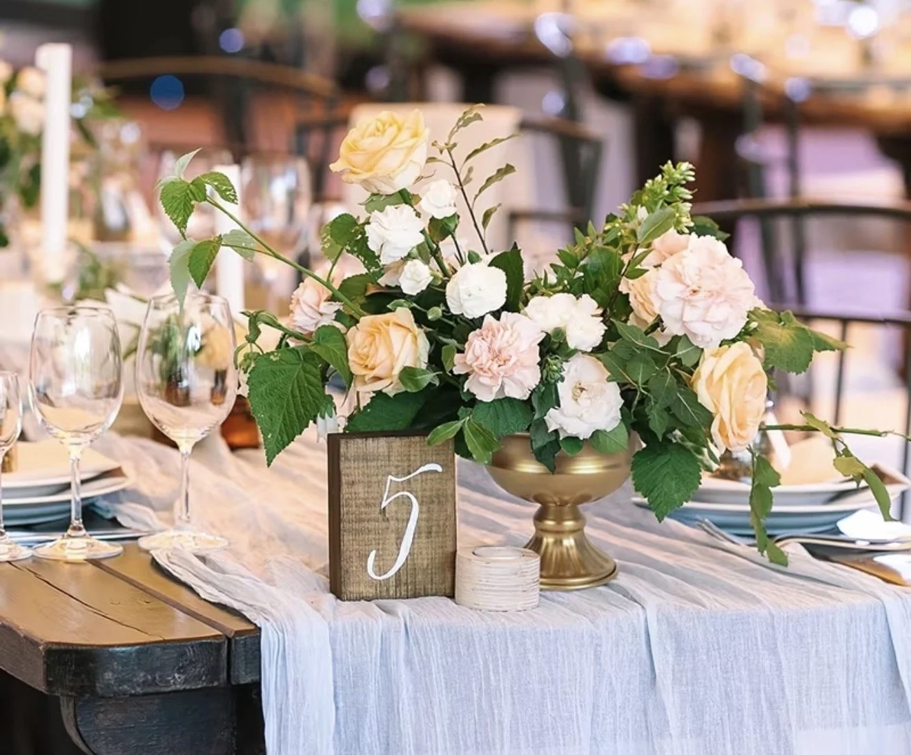 A table decorated with a centerpiece of peach and white roses, white hydrangeas, and green leaves. There are empty wine glasses, a wooden table number sign with the number 5, and a white table runner. The background shows blurred chairs and tables.