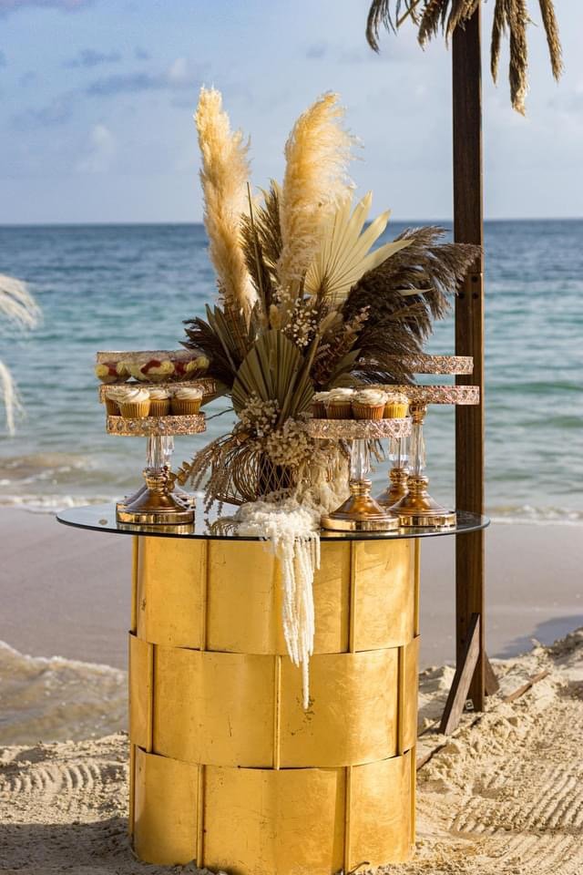 Decorative floral arrangement on a beachside table with cupcakes, set against ocean and sky background, under a palm tree.