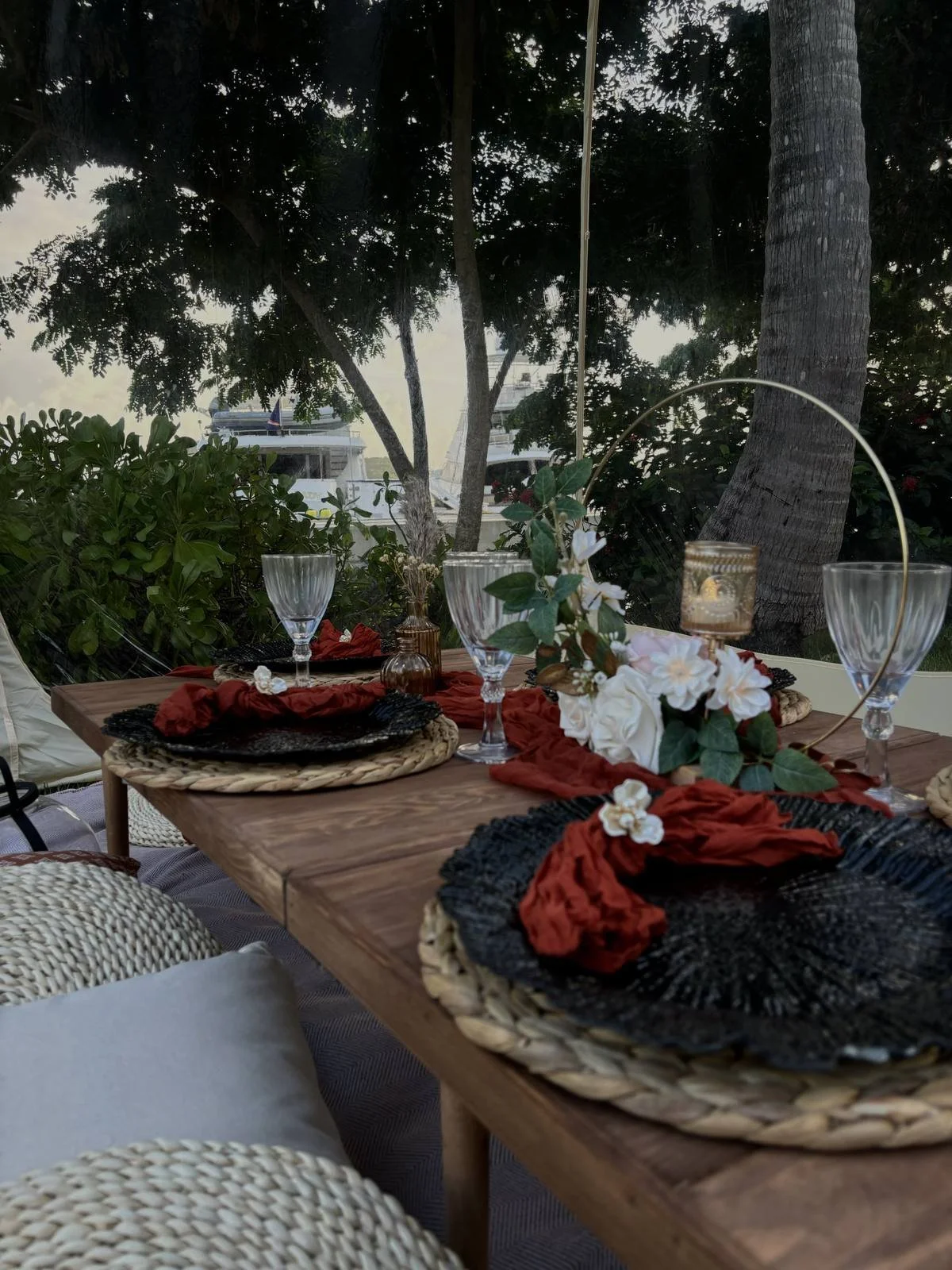 Outdoor dining table set with black woven placemats, red napkins, crystal glasses, and a floral centerpiece, with trees and a boat visible through a window in the background.