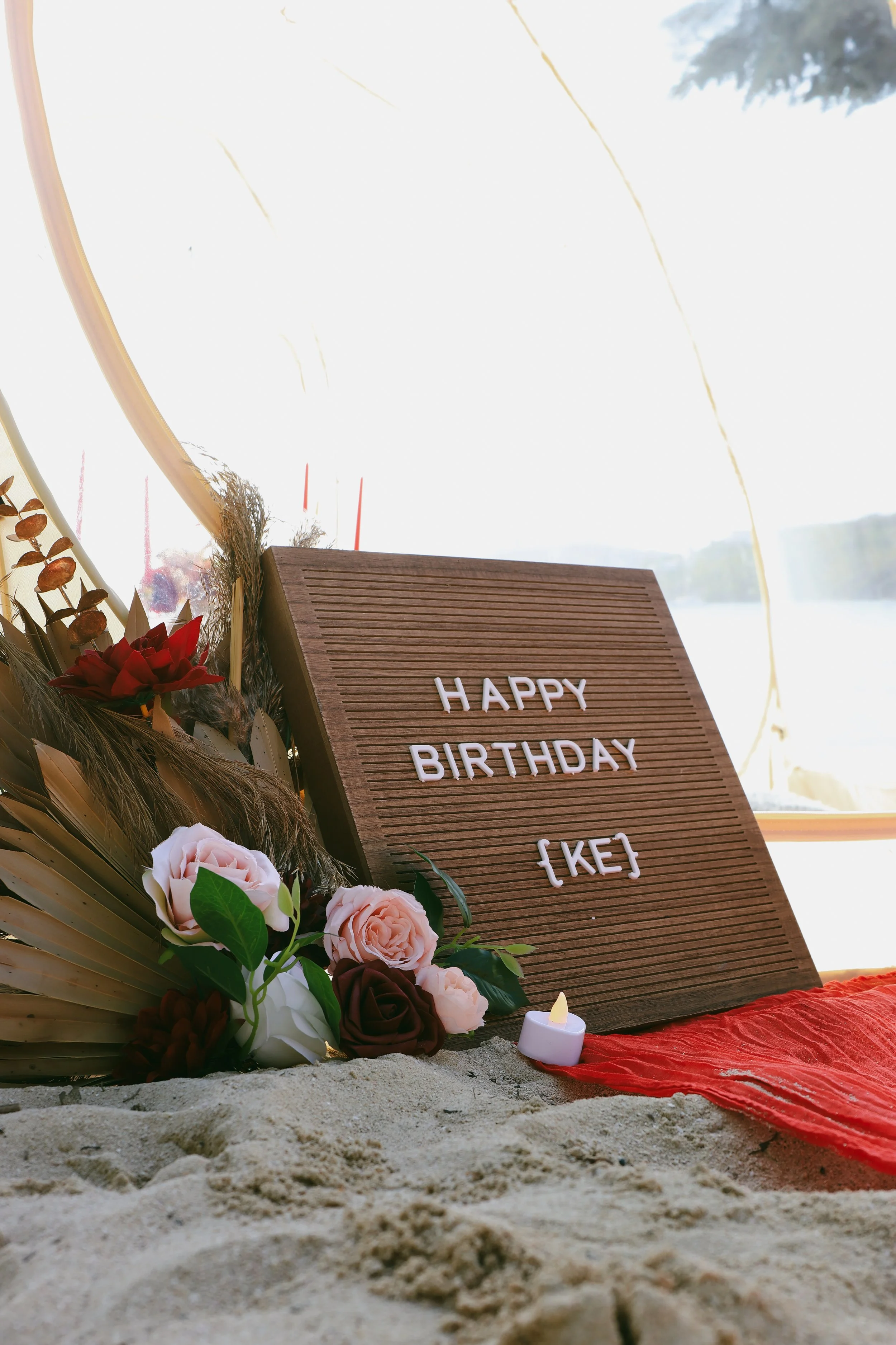 Beach scene with a wooden message board that says 'HAPPY BIRTHDAY [KEJ]'. Flowers and a small candle are placed around the board, and there are yellow balloons in the sky.