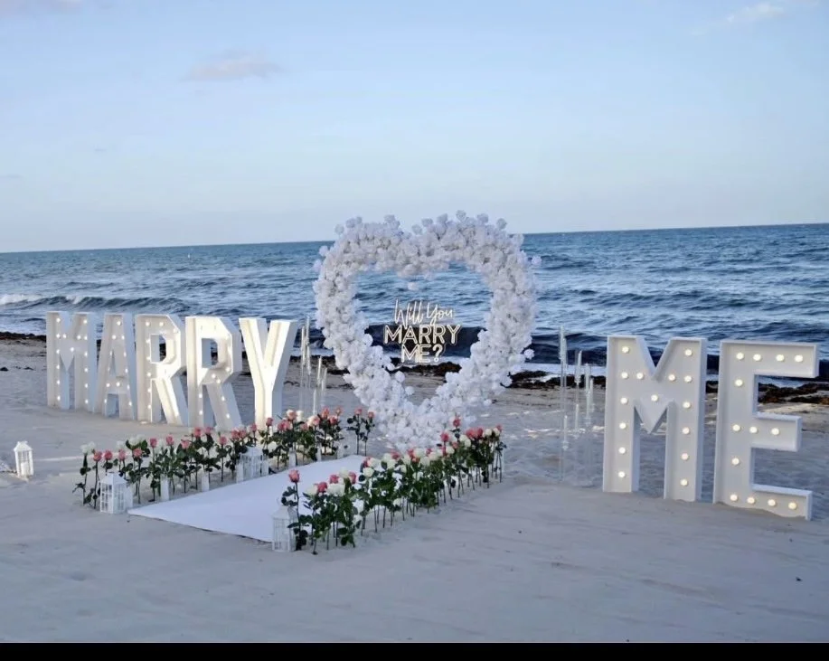 Wedding proposal setup on a beach with large illuminated 'MARRY ME' letters, a heart-shaped floral arrangement, and pink roses with lanterns surrounding the setup.