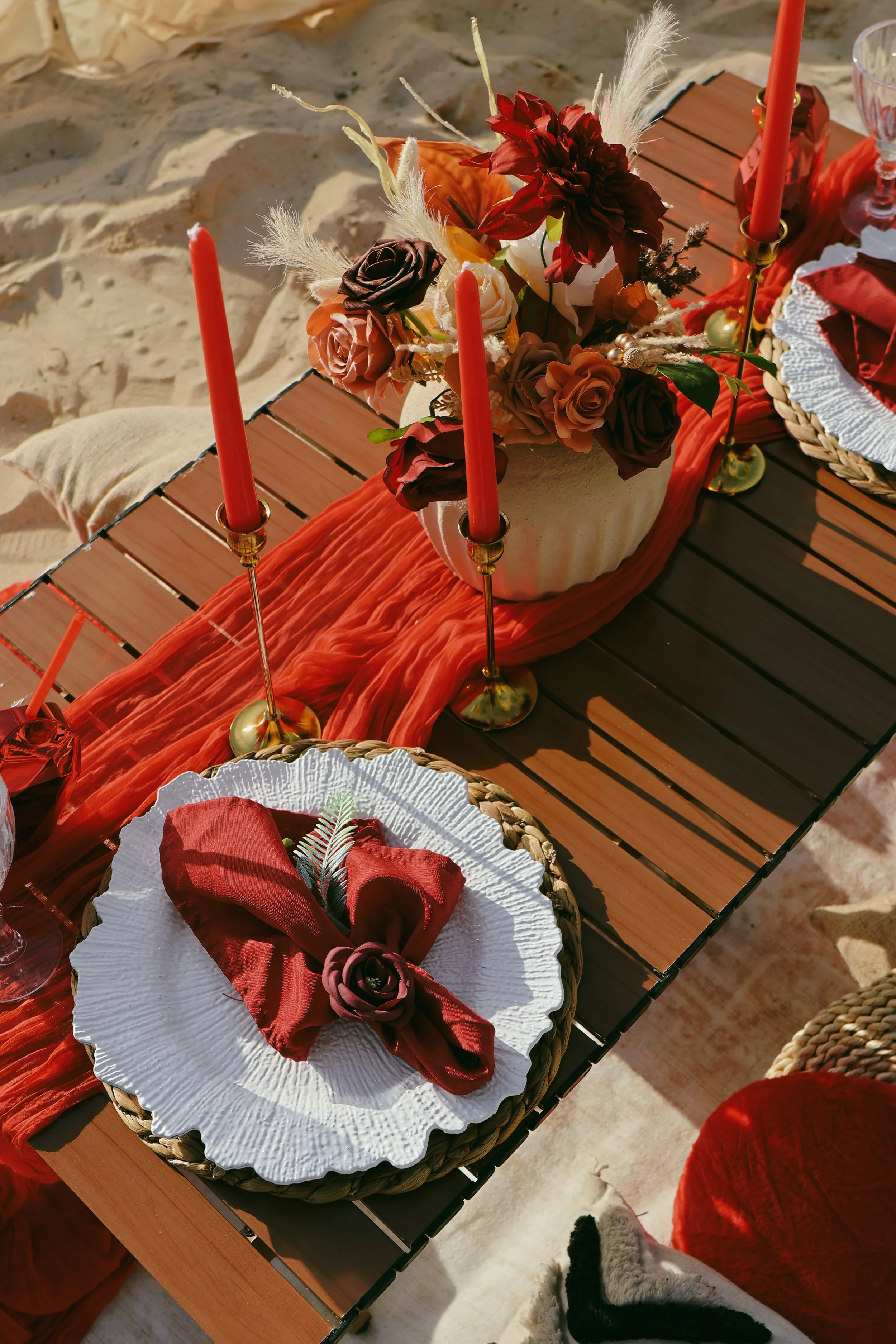 A festive table setup on a wooden table with a centerpiece of mixed dark red, light pink, and peach roses, accented with dried foliage and feathers in a white vase. The table is decorated with a red table runner, gold candlesticks with red candles, a