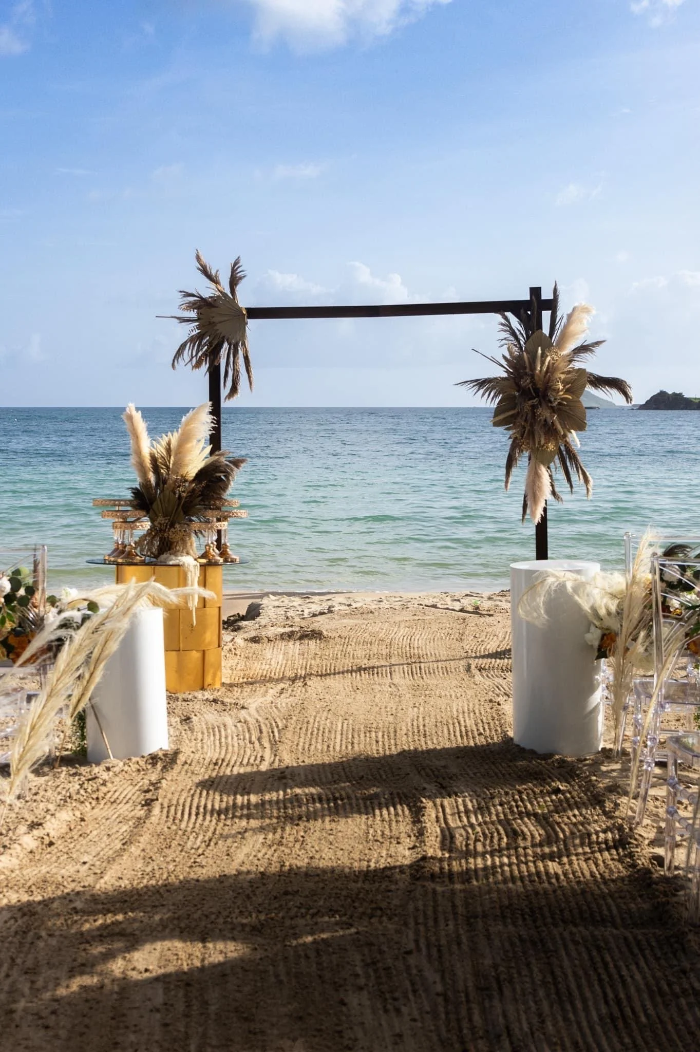 Beachside wedding setup with an arch decorated with dried palm leaves and pampas grass, white candles, and chairs with transparent backs, overlooking the ocean.