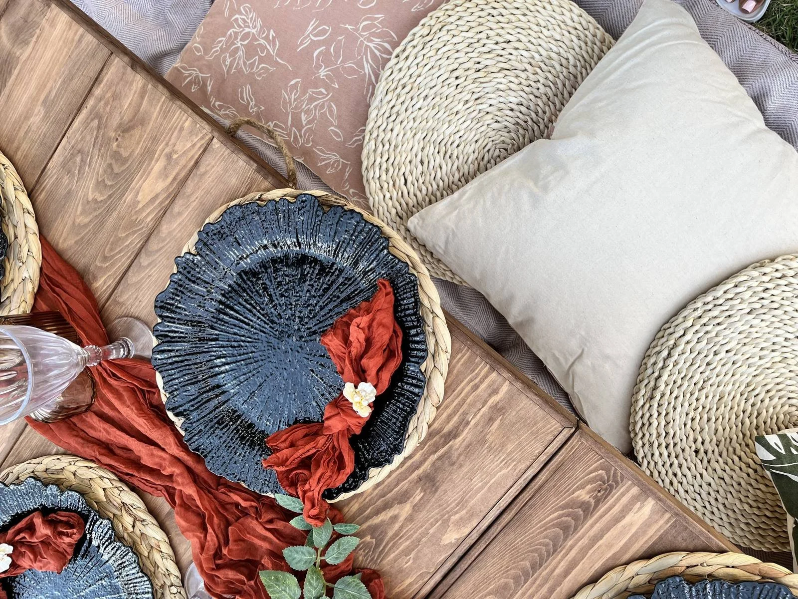 Top-down view of a table setting with woven placemats, cushions, and decorative plates, including a black ceramic plate with a red cloth and small white flower, set on a wooden table with a glass and cloth napkins.