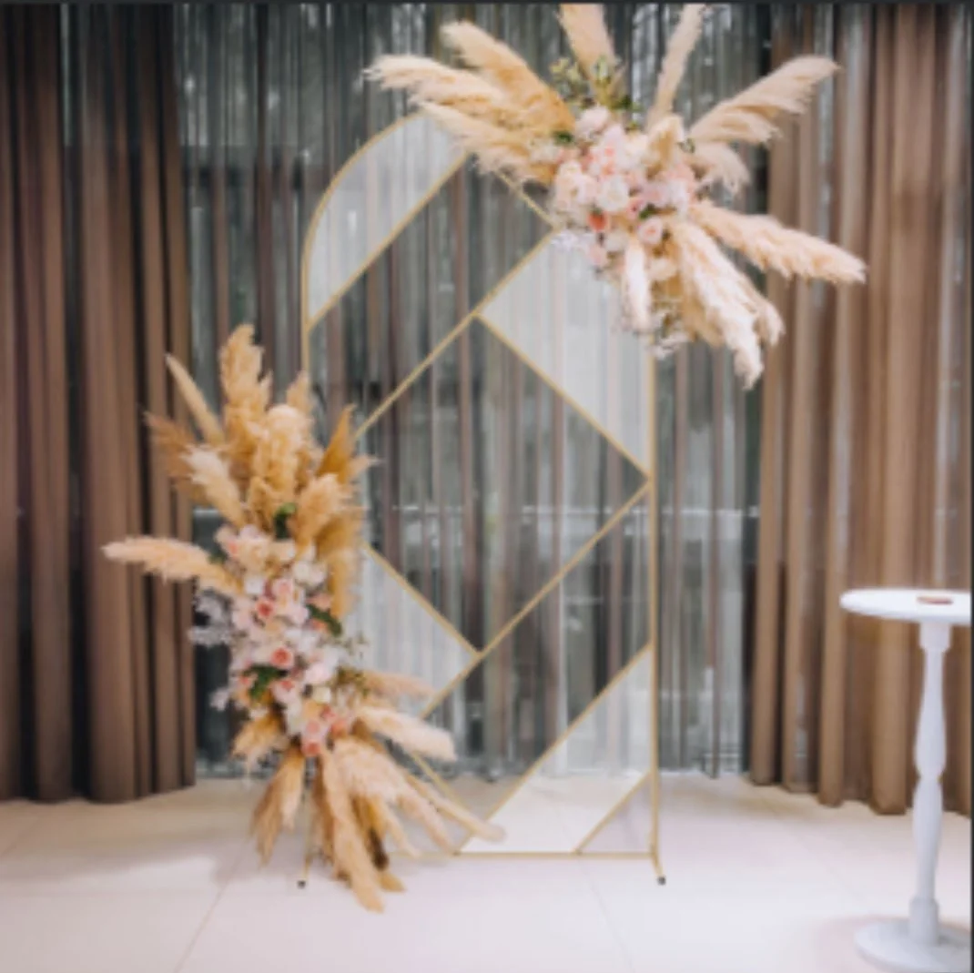 Decorative floral arrangement with pink and cream flowers and pampas grass on a gold geometric frame in front of a window with brown curtains.