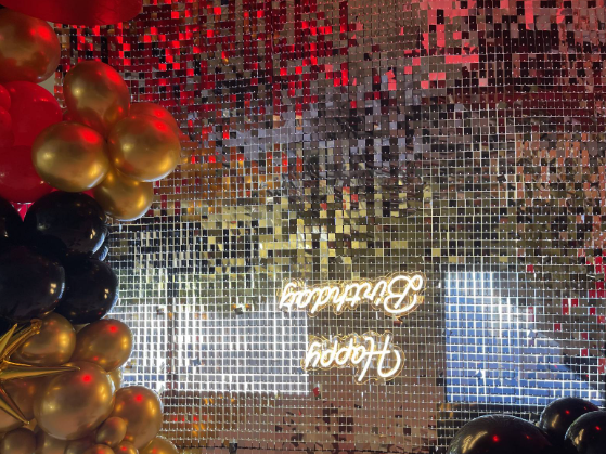 Decorative backdrop featuring a mirror surface covered with small square tiles, with a balloon arrangement of gold, black, and red balloons on the left and bottom sides, and a neon sign that reads "Happy Birthday" in cursive on the mirror.