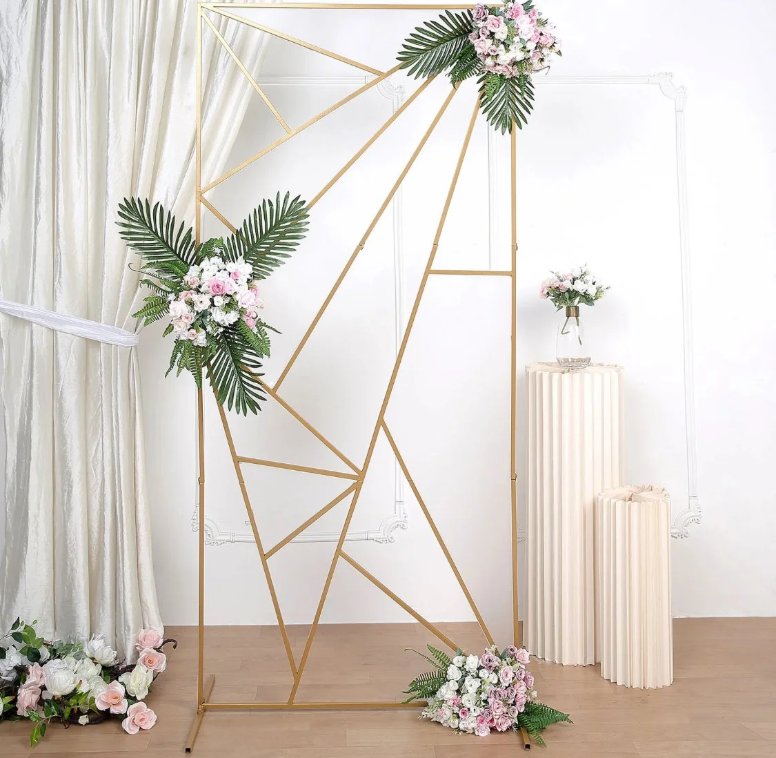Decorative geometric gold frame with floral arrangements of pink and white roses, green leaves, and orchids, set against white curtains and pedestal stands with matching vases of flowers.