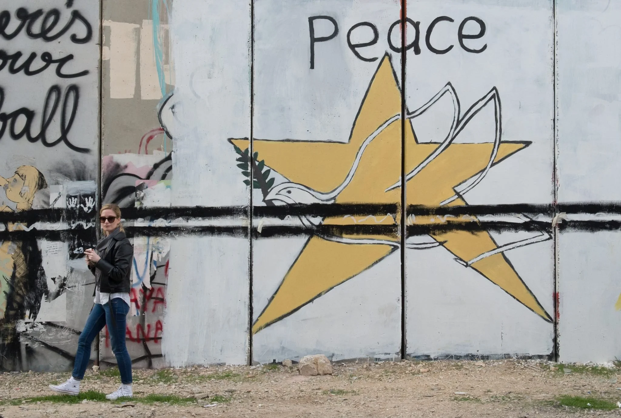 A woman with sunglasses wearing a black leather jacket, white shirt, and jeans walking past a large mural on a wall featuring a star, a dove, and the word "peace" written above.