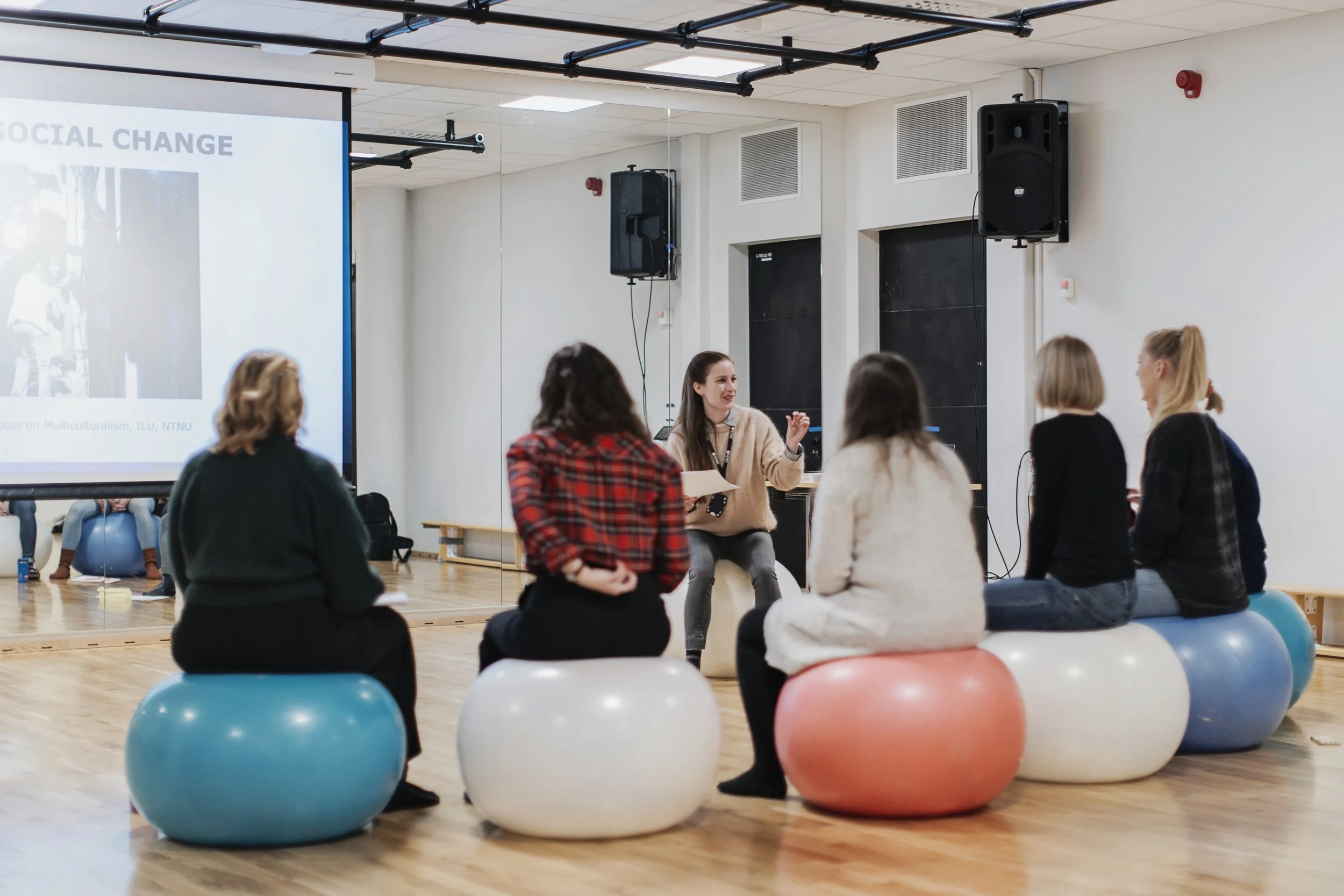 A woman leading a discussion or presentation to a group of people seated on exercise balls in a room with a large mirror and a projection screen.