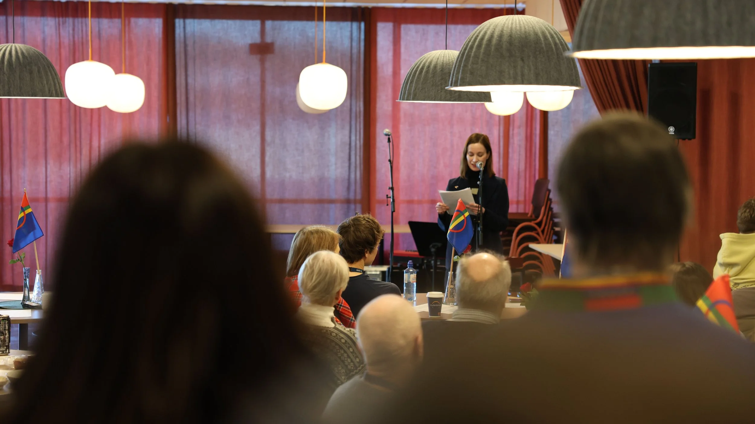 A woman speaking at a conference or event, standing behind a microphone and reading from papers, with an audience seated in front of her. The room has large hanging lights and red curtains, with small flags on tables.