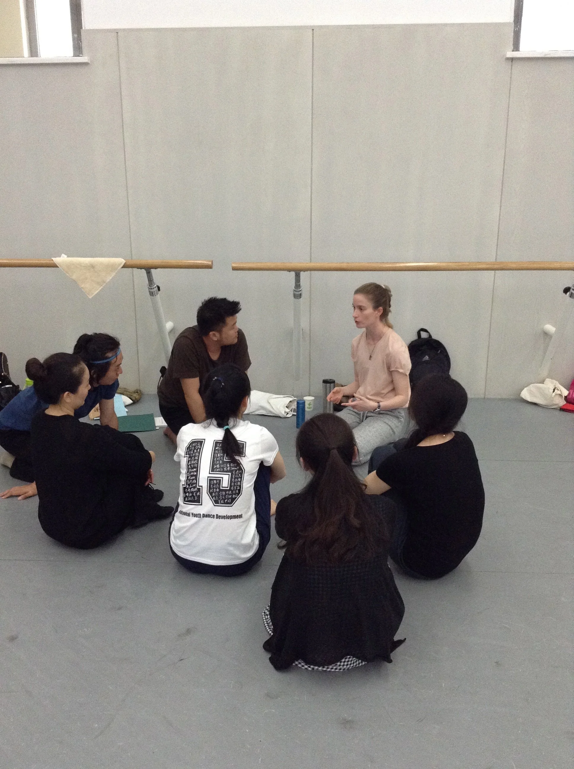 A group of young people, sitting on the floor, listening to a woman speaking in a dance studio with ballet barres and grey walls.
