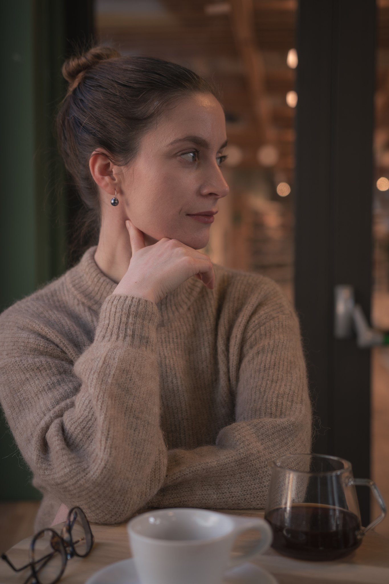A woman with brown hair tied in a bun, wearing a beige sweater and black earrings, sits at a table with coffee cups and glasses, looking thoughtfully to the side inside a cozy, warmly lit cafe with wood accents in the background.