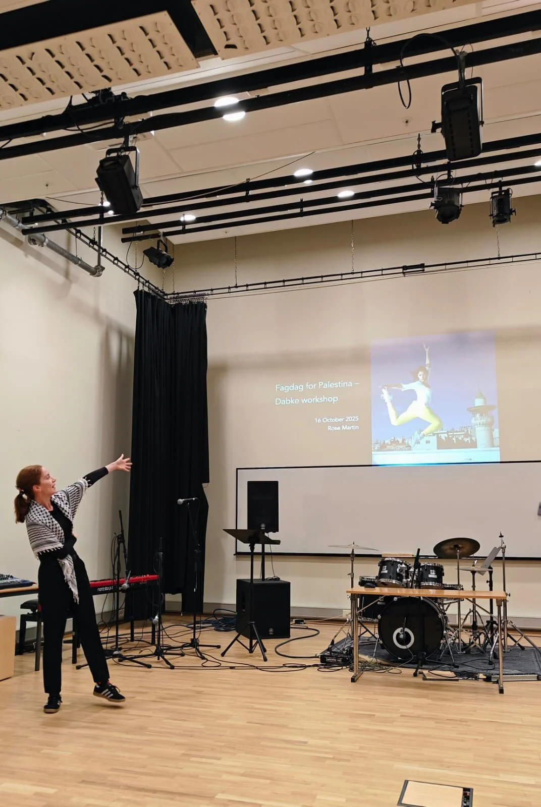A woman is standing in a classroom or studio, pointing towards a screen on the wall. The screen displays a presentation titled 'Fagdag for Palestina - Dabble workshop' dated October 16, 2025, with the name Rose Martin. There is musical equipment, including a drum set, microphone, and music stand, set up in front of the screen. A black curtain is partially drawn to the side.