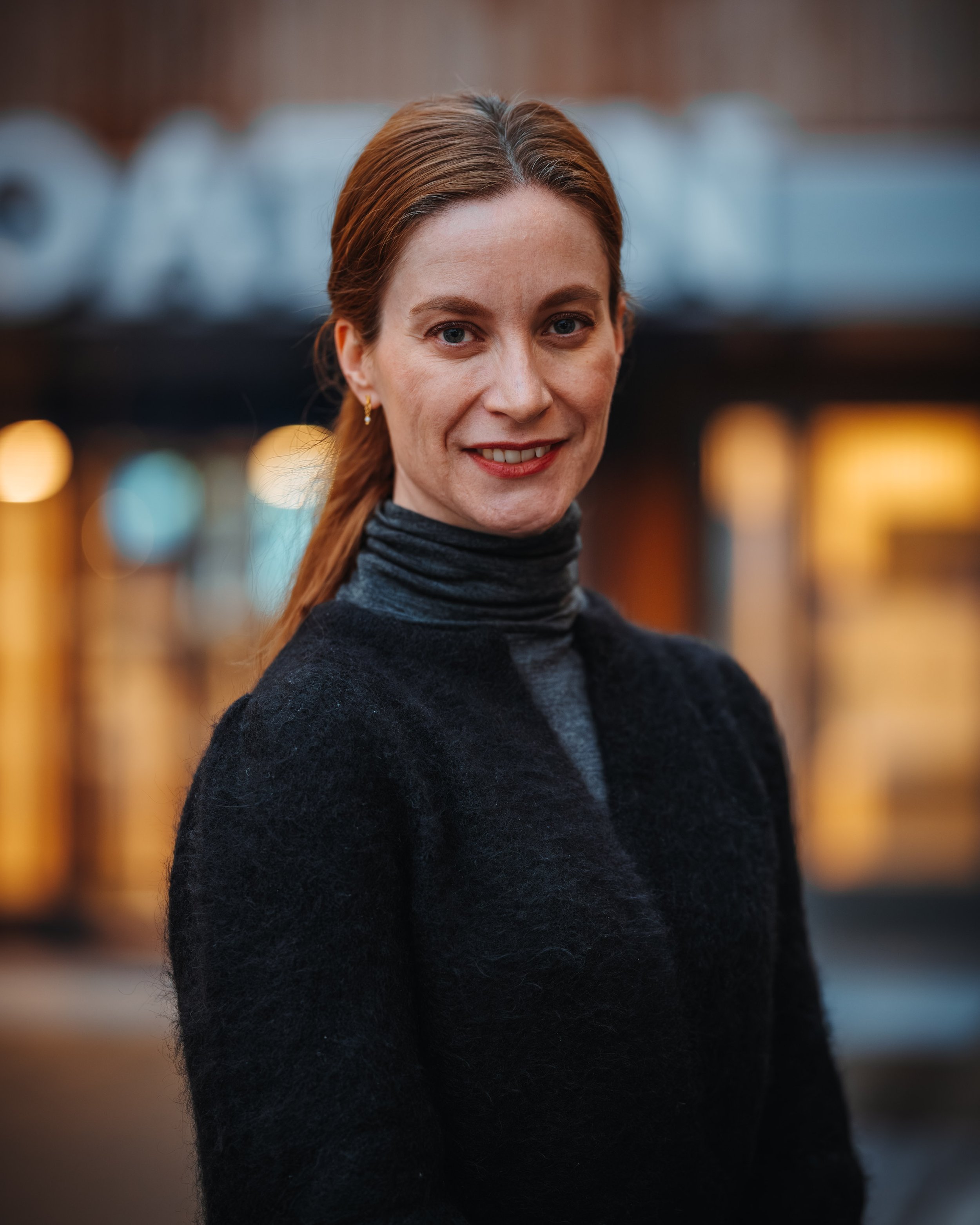 A woman with red hair in a ponytail, wearing earrings and a dark turtleneck, smiling in an indoor setting with blurred lights in the background.
