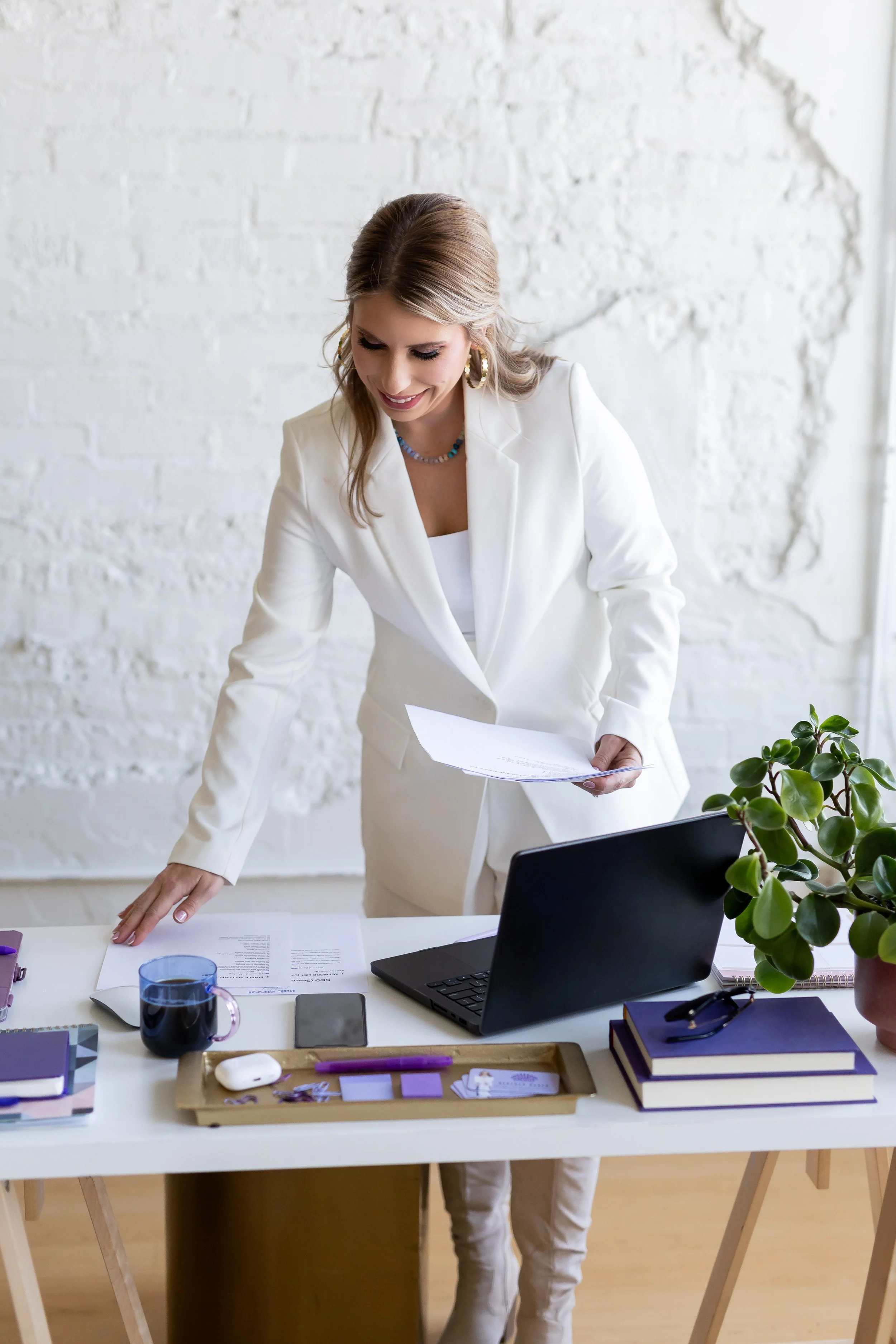 A woman in a white suit standing at a desk, looking at papers and smiling, in an office with a white brick wall and a potted plant.