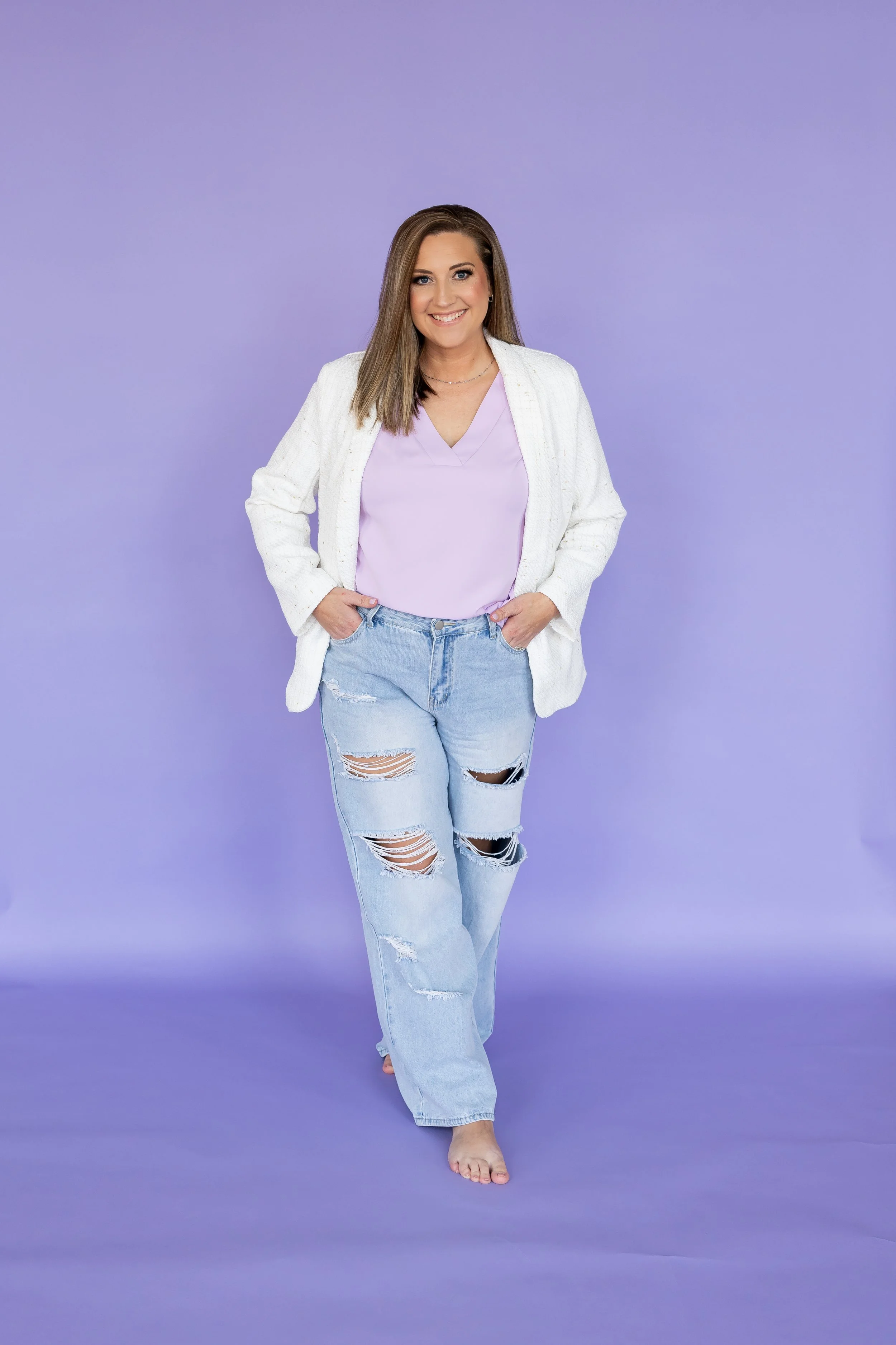 Woman with long brown hair smiling, wearing a white blazer, lavender top, and ripped jeans, walking barefoot against a purple background.