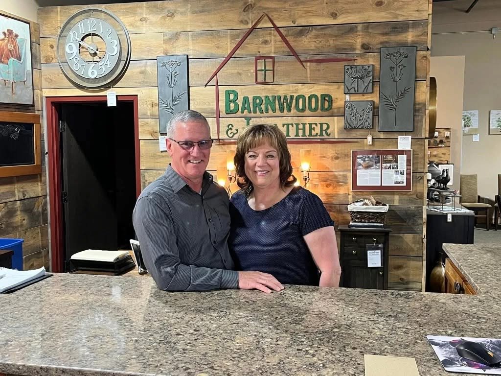 A smiling man and woman standing behind a granite reception counter inside a rustic store or office, with wall art and a clock in the background.