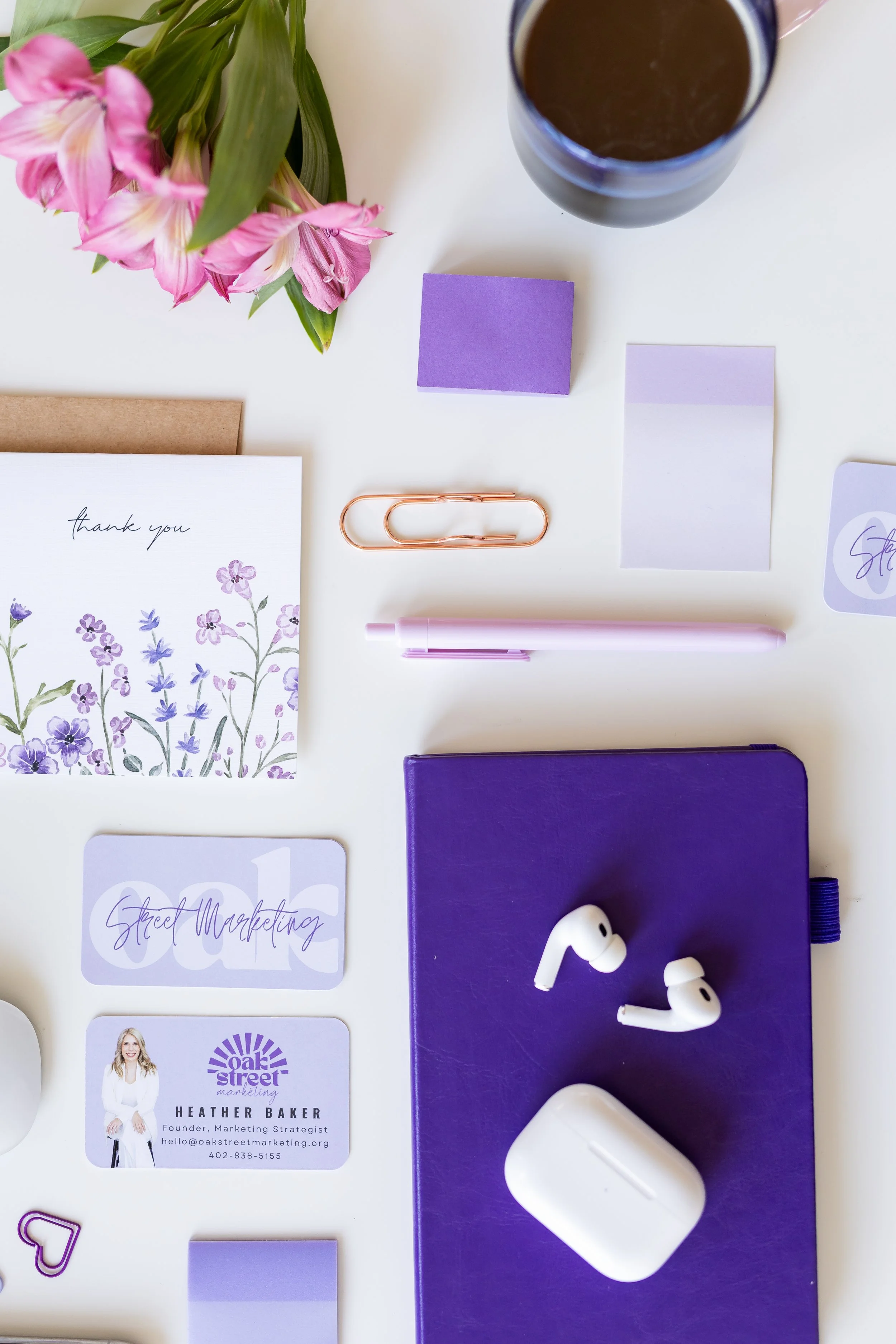 A flat lay of office supplies and personal items on a white surface, including pink flowers, a glass of coffee, purple sticky notes, a thank you note with purple floral design, business cards, a purple notebook, white wireless earbuds, a white AirPods case, a white pen, a purple paper clip, and a purple folder.