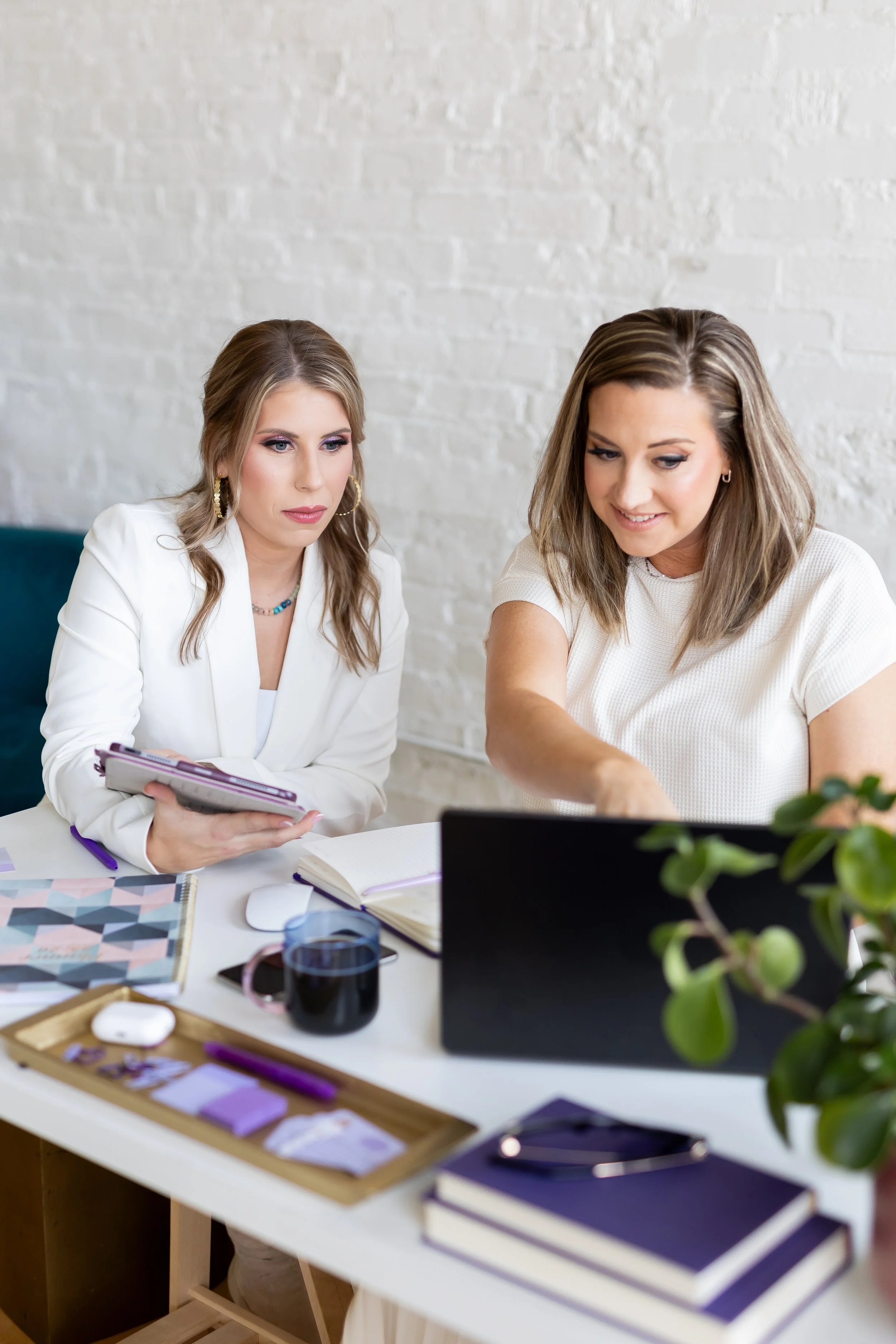 Two women sitting at a desk working on computers with notebooks, books, pens, and a cup of coffee visible on the desk, against a white brick wall background.