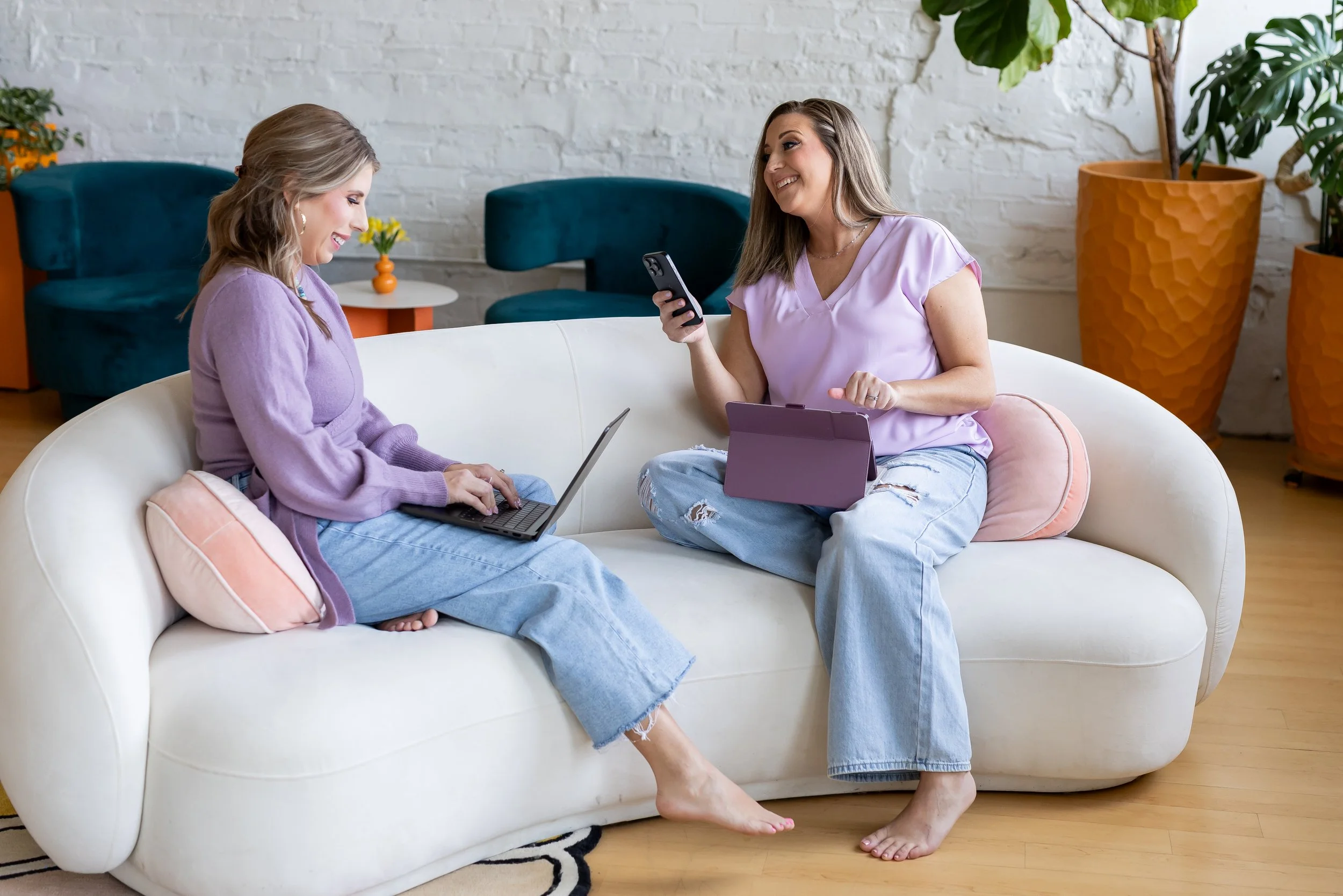 Two women sitting on a white couch in a brightly lit room, smiling and engaging with each other. One woman is using a laptop, and the other is holding a smartphone with a purple case, resting on her lap. The room has white brick walls, green plants, and minimalist decor.
