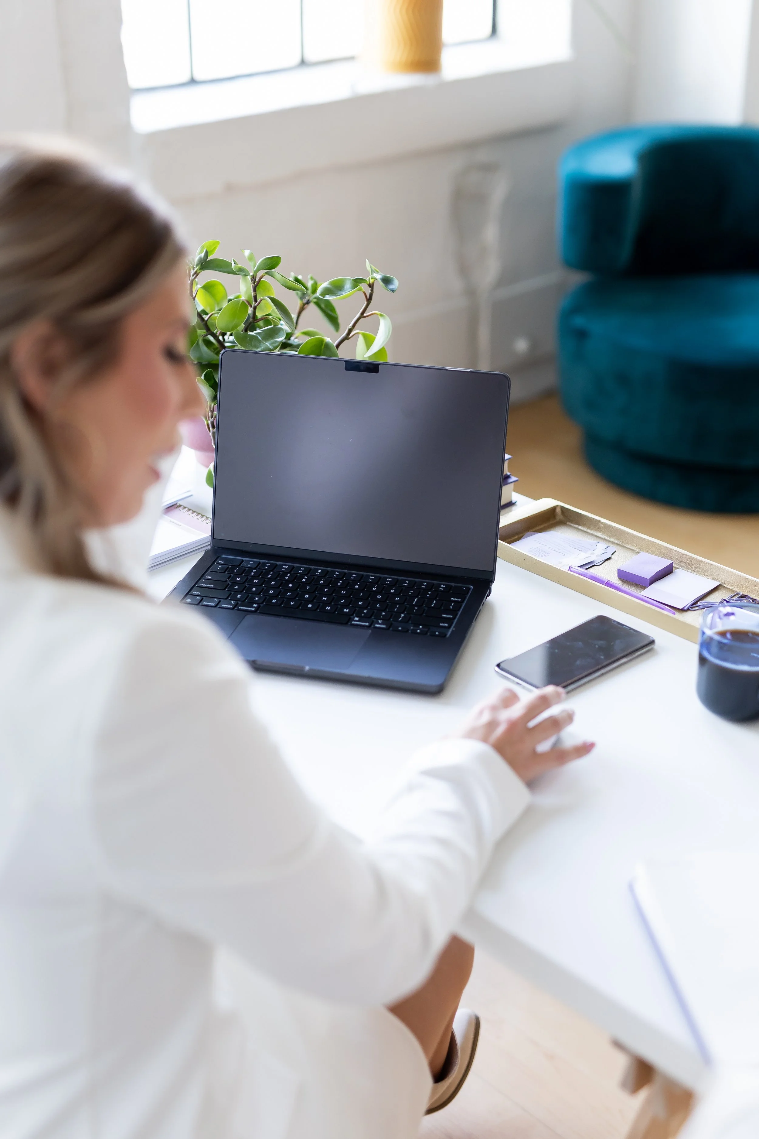 A woman with blonde hair working at a white table in a bright room, with a closed laptop, a tray of stationery, a smartphone, and a black mug. In the background, there is a window, a yellow vase, a green plant, and a teal chair.