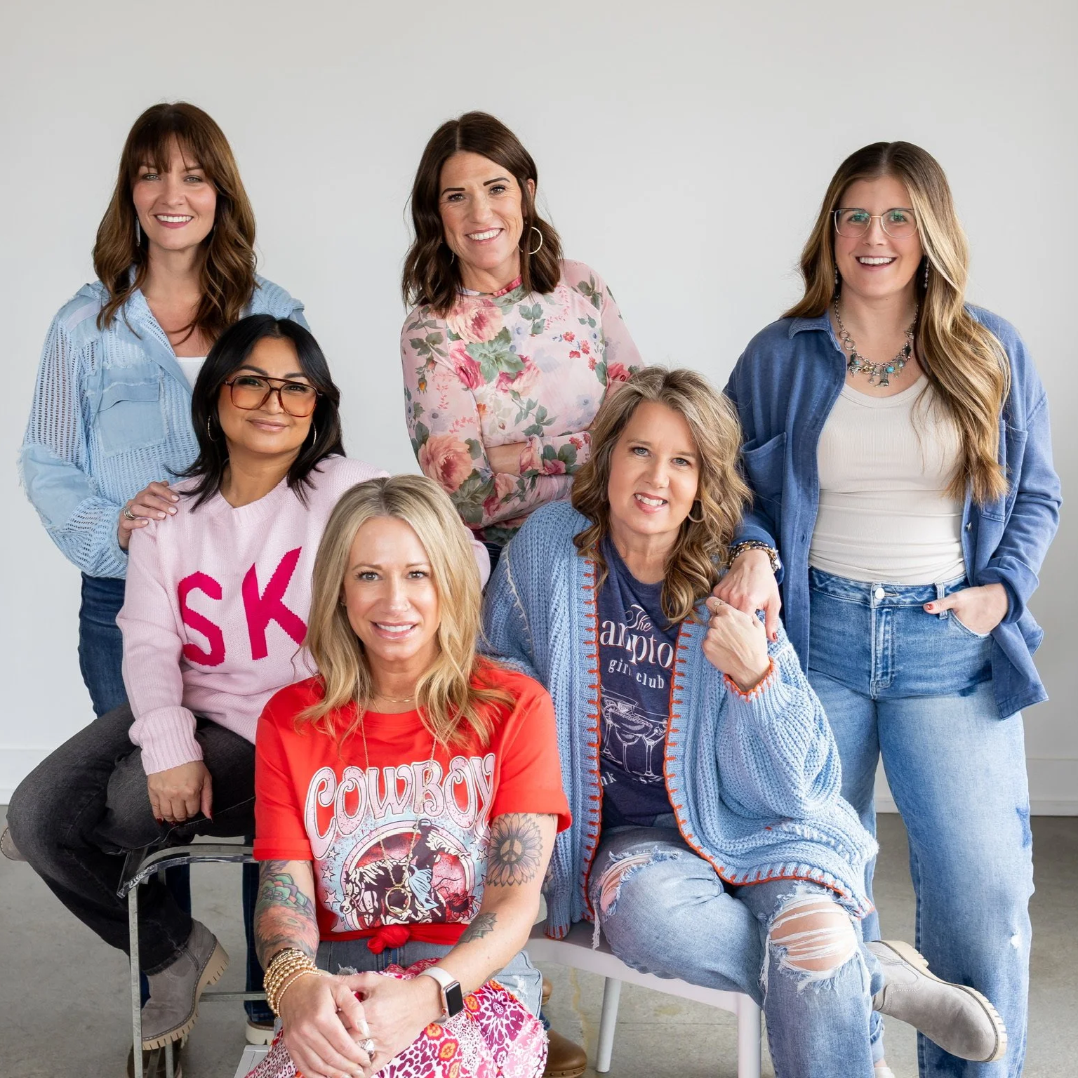 A group of seven women posing together indoors against a plain white background, smiling at the camera. They are dressed casually in colorful and patterned clothing.
