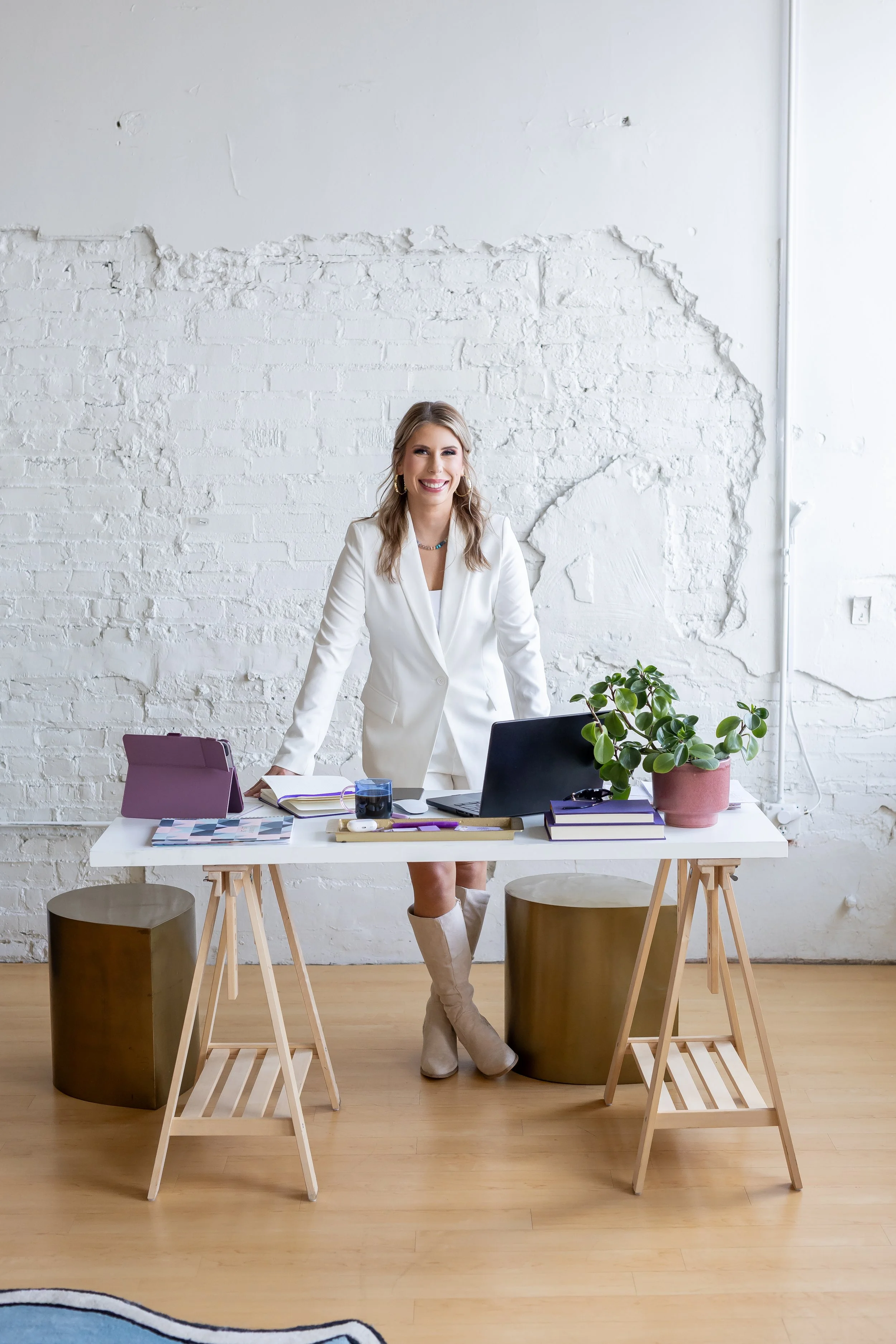 A woman standing at a white office desk filled with notebooks, a laptop, a tablet, a glass of water, and a potted plant. She is wearing a white blazer and boots, smiling, in front of a white brick wall.