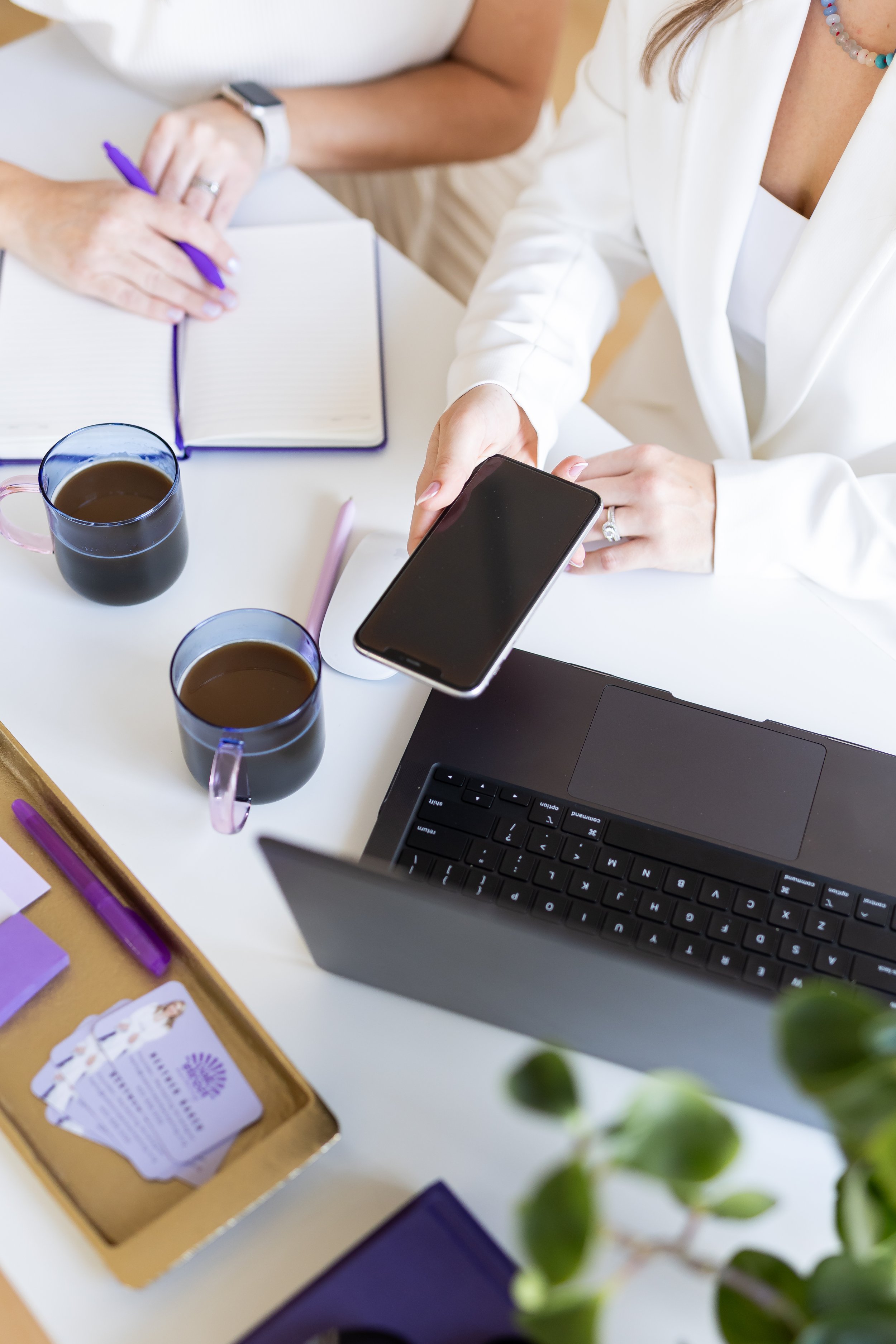 Two women working at a desk with notebooks, three cups of coffee, a laptop, a smartphone, and some colorful markers, with a plant partially visible in the foreground.