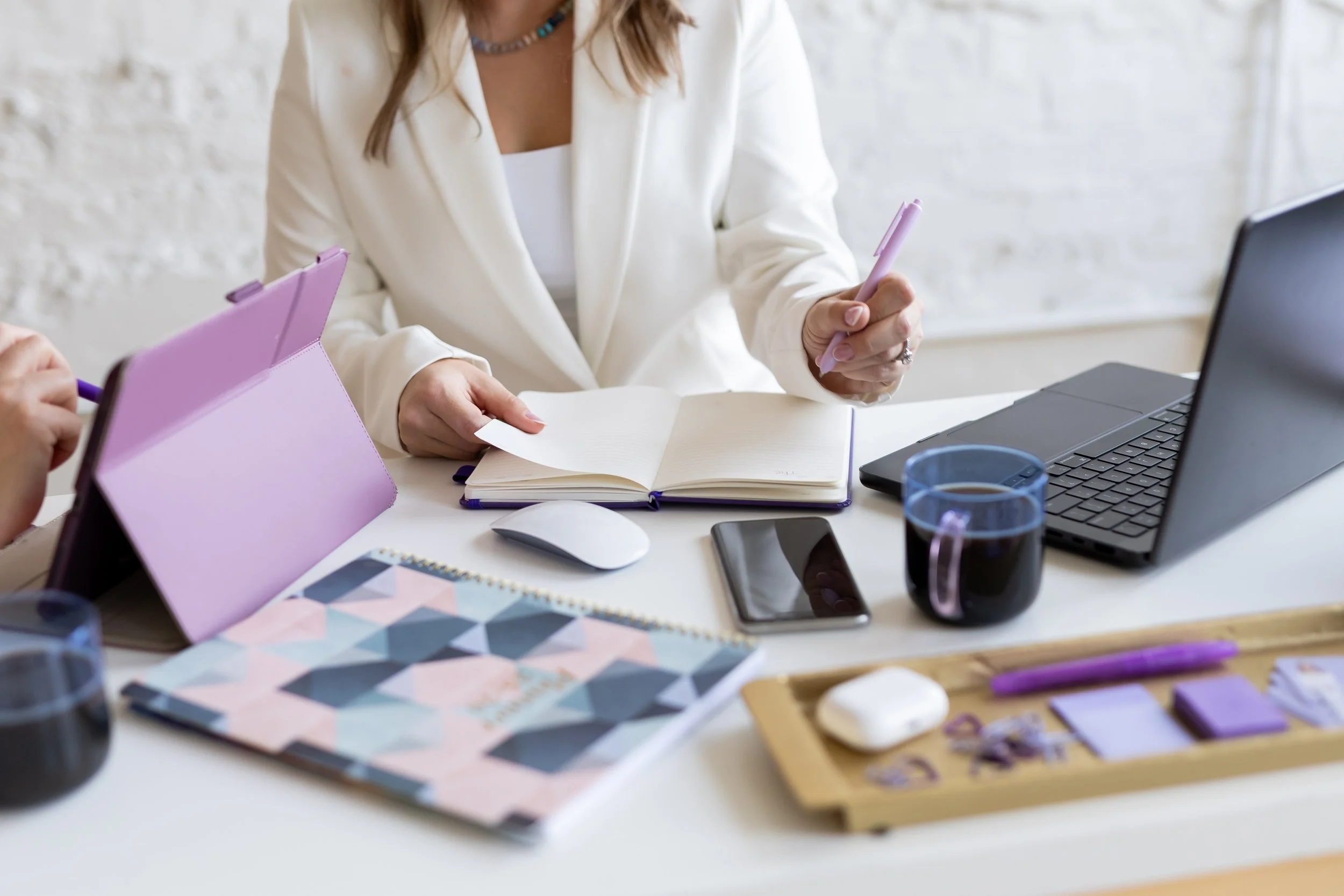 A woman in a white blazer working at a desk with a laptop, notebooks, pens, purple tablet, and glasses of water.
