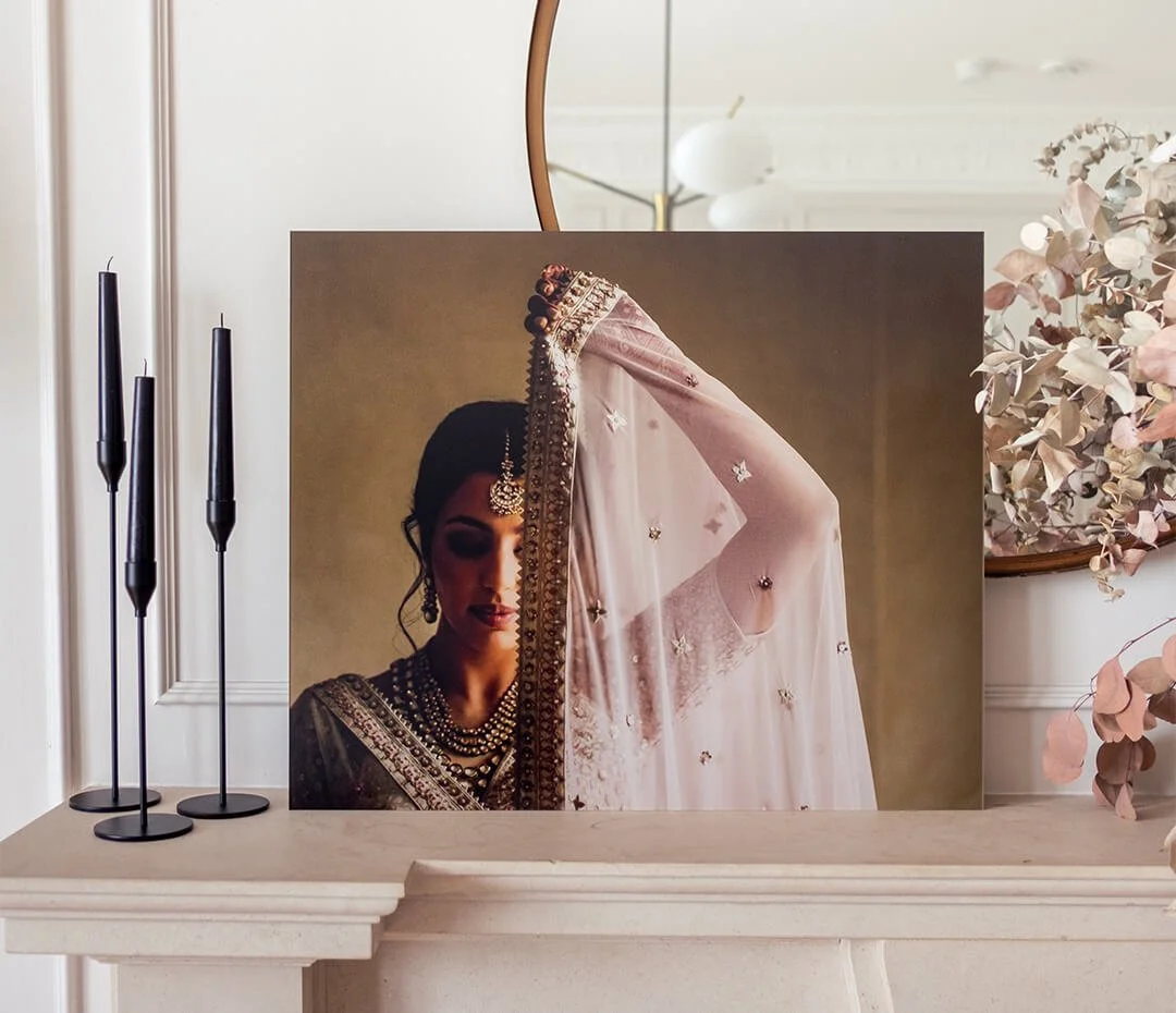 A decorative canvas print of a woman in traditional Indian attire, jewelry, and a veil covering her face, placed on a fireplace mantel with black candles and pink floral decor surrounding it.