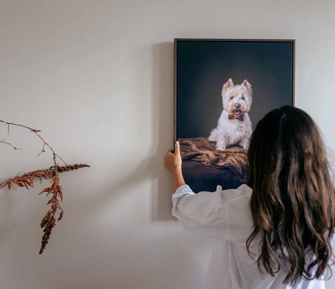 A girl with long wavy hair is holding up a framed portrait of a cute white dog with tan markings, sitting on a blanket, against a dark background.