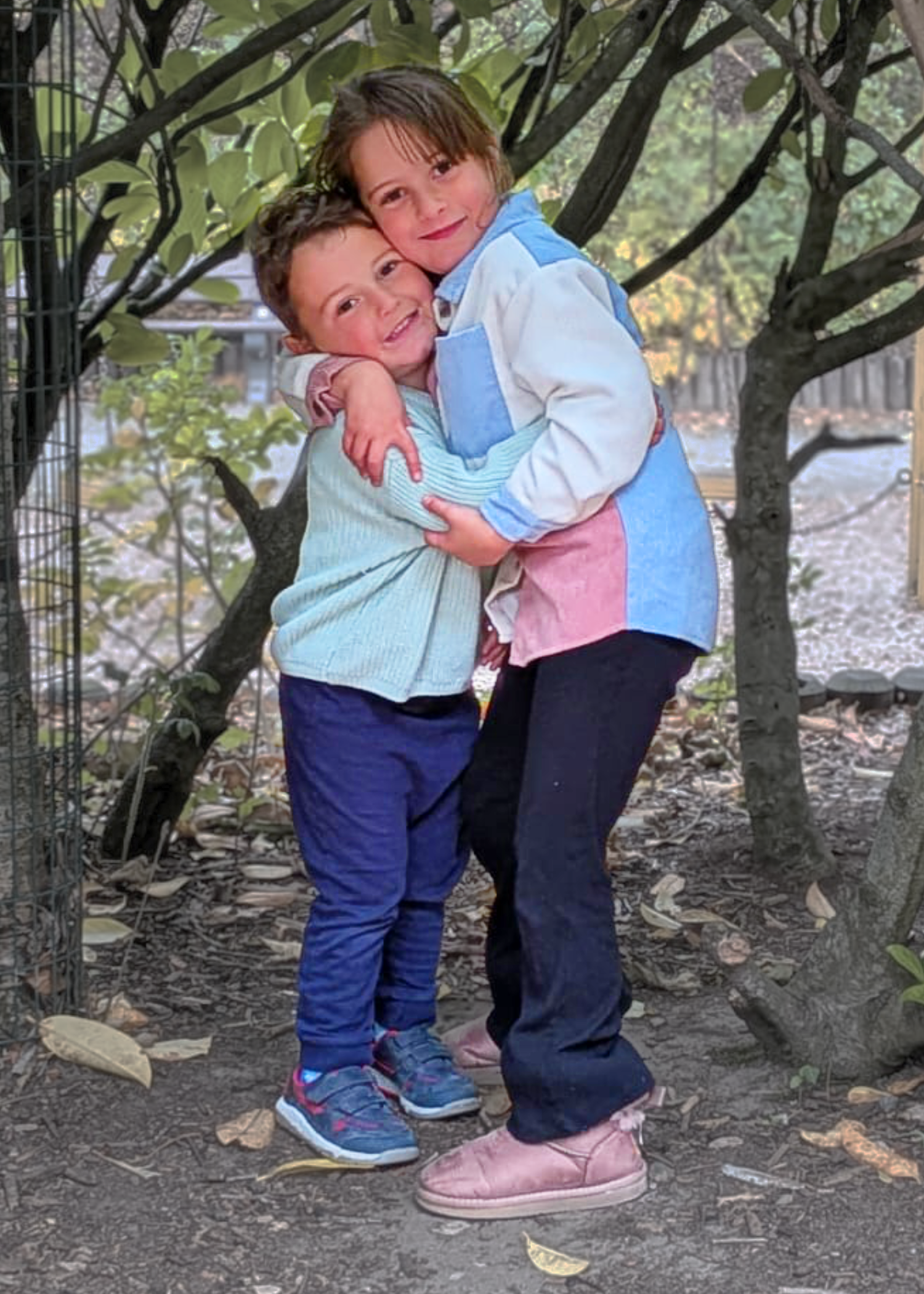 Two children, a boy and a girl, hugging each other and standing outside on a path, surrounded by trees with leaves on the ground.