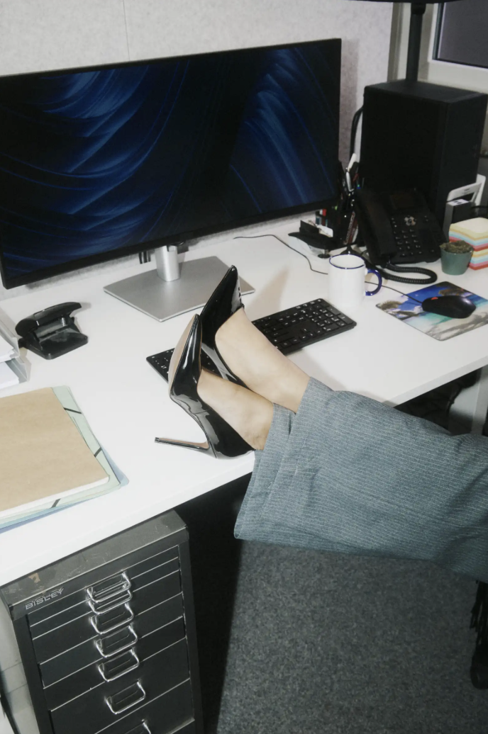 Office desk with a large monitor, keyboard, mouse, and office supplies. Person is crossing their legs on the desk, wearing gray pants and black high-heeled shoes.
