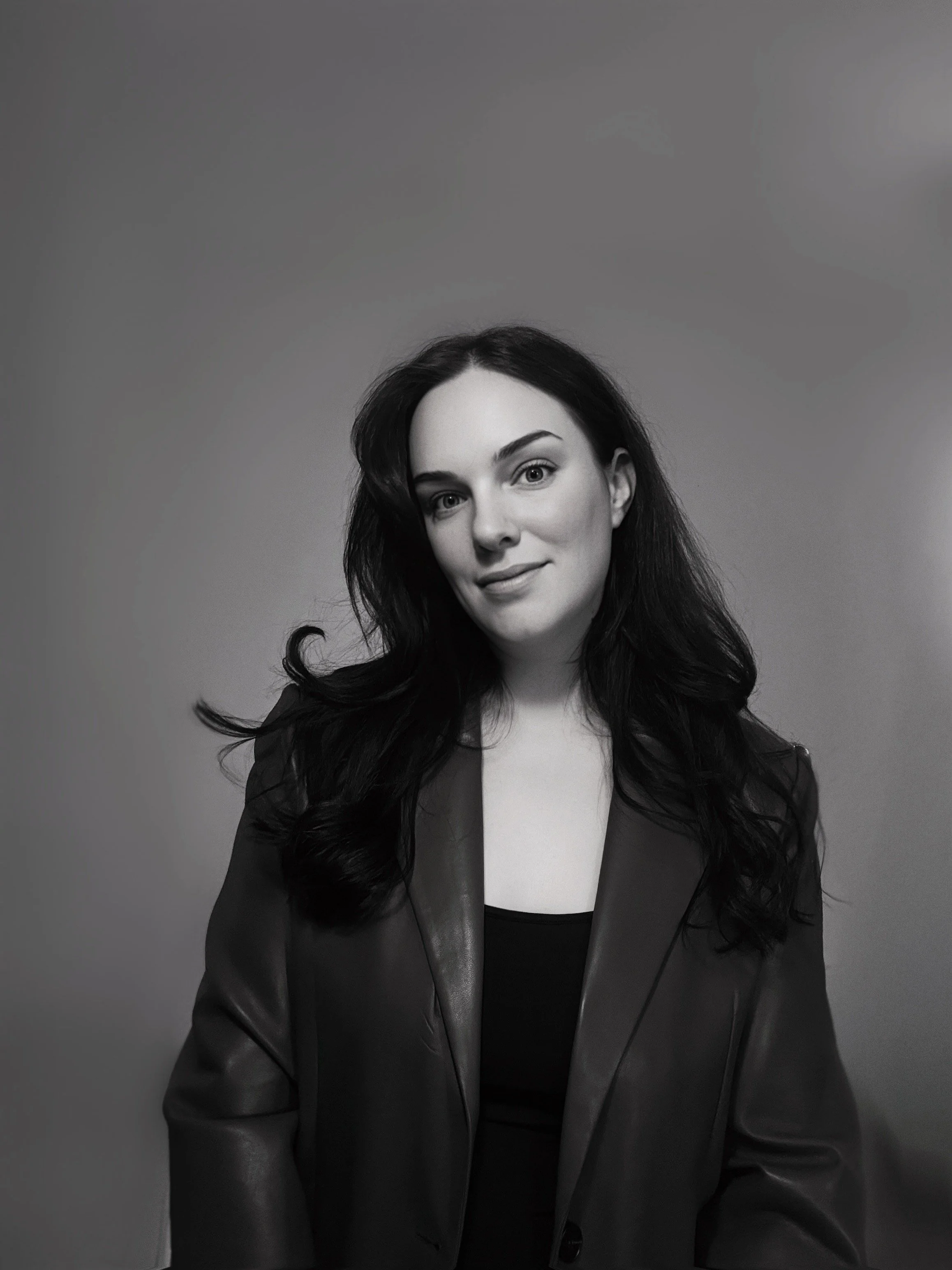 Black and white portrait of a woman with long wavy dark hair, wearing a dark blazer and a black top, against a plain background.
