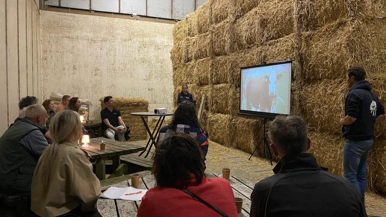 Een groep mensen zit in een hooischuur, kijkt naar een presentatie op een scherm dat een koe afbeeldt. Er staat een persoon voor het scherm die de presentatie geeft.
