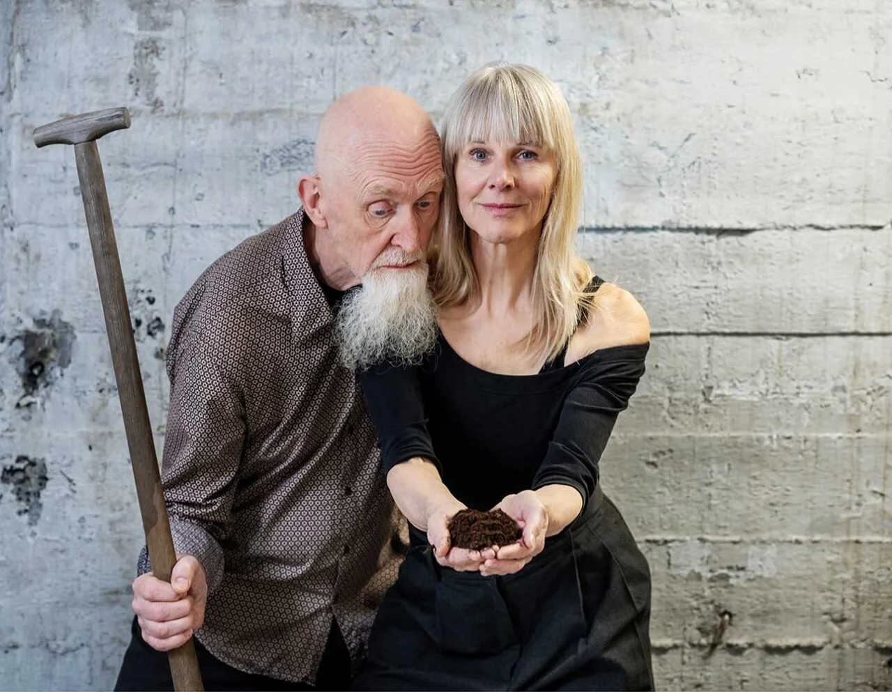 Een oude man met een witte baard en een jonge vrouw met blond haar die samen een stuk aarde vasthouden, tegen een lelijke, witte, gestuukte muur.