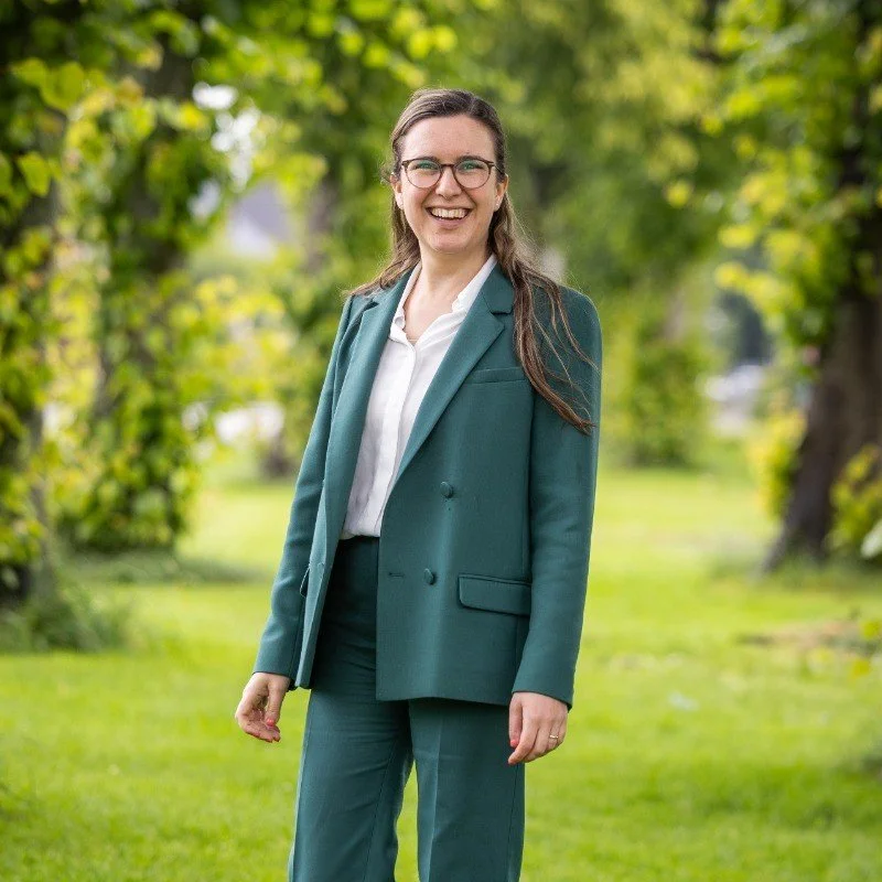 Vrouw gekleed in een groene blazer en broek, lachend in een groene tuin met bomen.